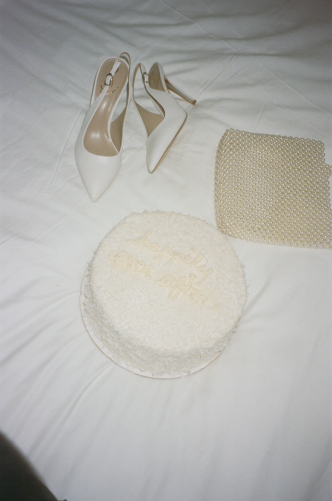 White high-heeled shoes, pearl clutch, and a cake with 'happily ever after' written on it, placed on a white surface.