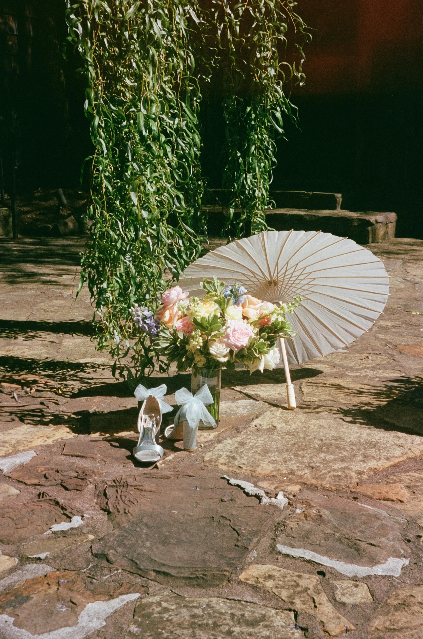 A bouquet of flowers with pink and white roses, a white paper parasol, and a pair of white high-heeled shoes with bows are arranged on a stone pavement next to hanging green foliage.