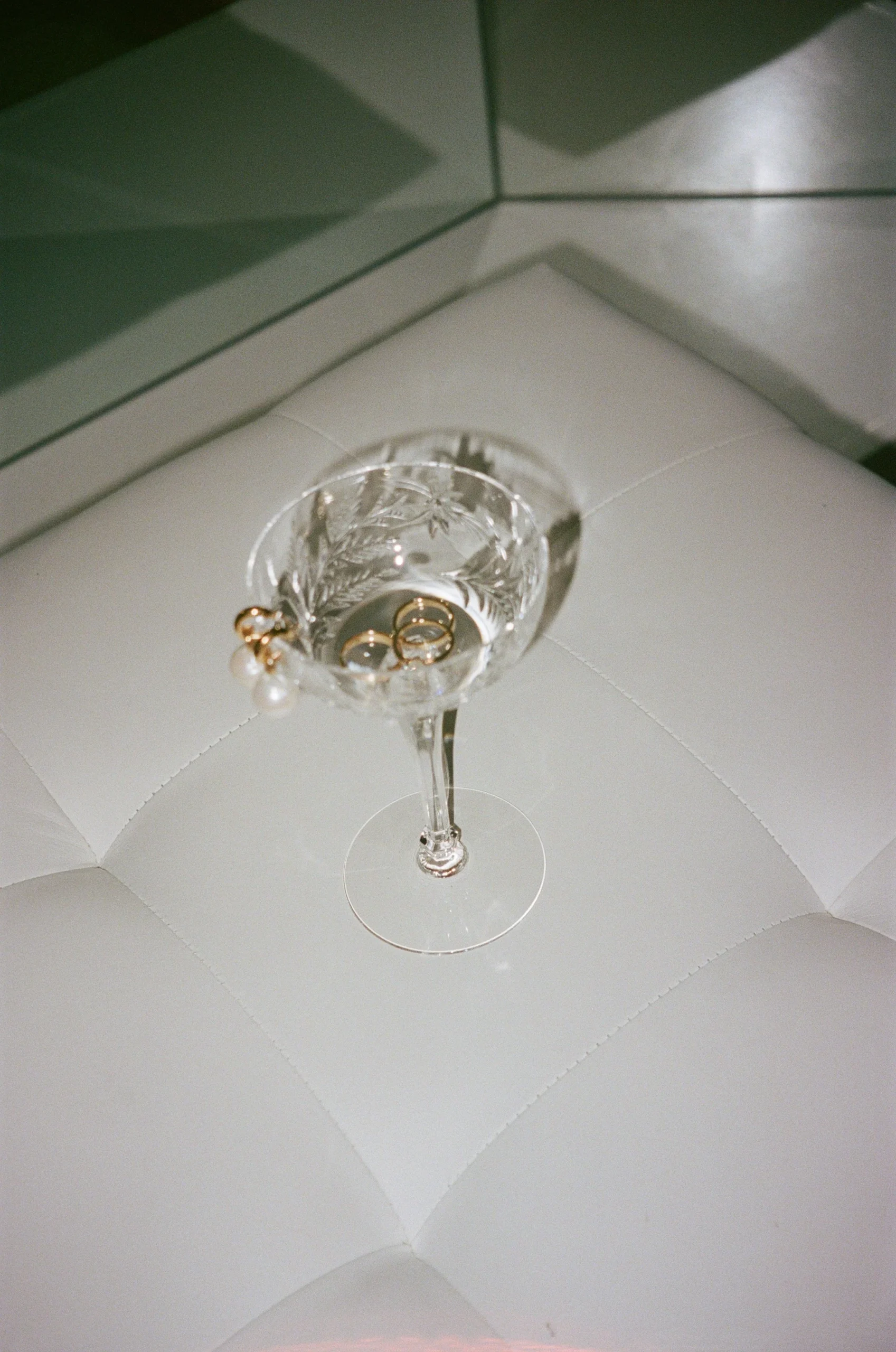 Crystal glass with water, holding two gold rings and pearl earrings, on a white tufted surface.