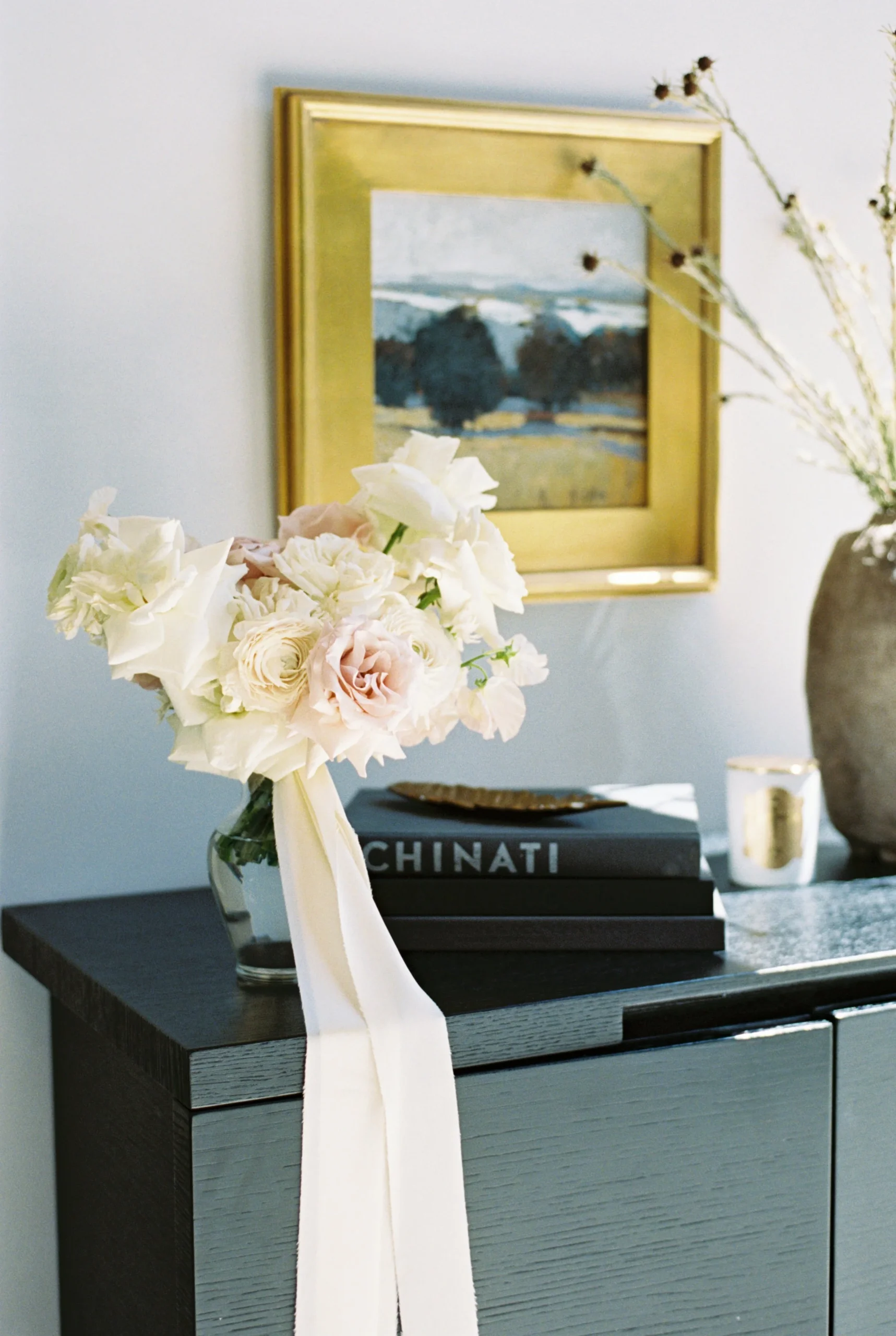 A bouquet of white and light pink flowers in a glass vase with a ribbon, placed on a dark wooden cabinet next to stacked books and a small gold dish. A framed painting hangs on the wall above.
