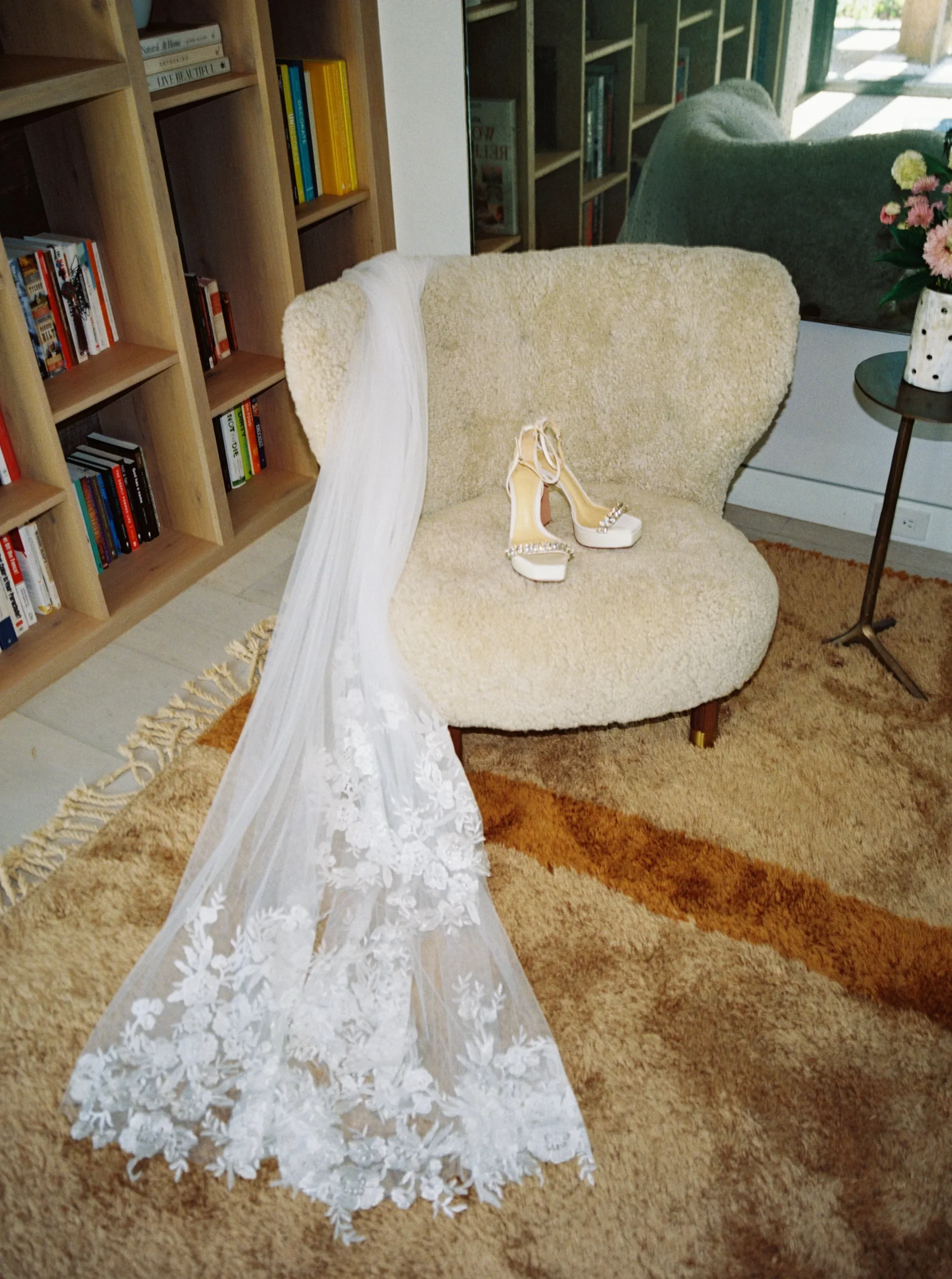 A beige upholstered chair with a pair of elegant high-heeled shoes on it. A white floral lace veil is draped over the chair. The setup is in a home interior with a bookshelf, a mirror, and a side table with a vase of pink flowers. A textured rug cove