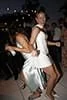 Two women dancing at an outdoor event at night, wearing elegant dresses, with decorative lights and people in the background.