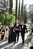 Three elegantly dressed people walking down an outdoor aisle, flanked by tall trees and guests seated on either side, in a wedding or formal event setting.