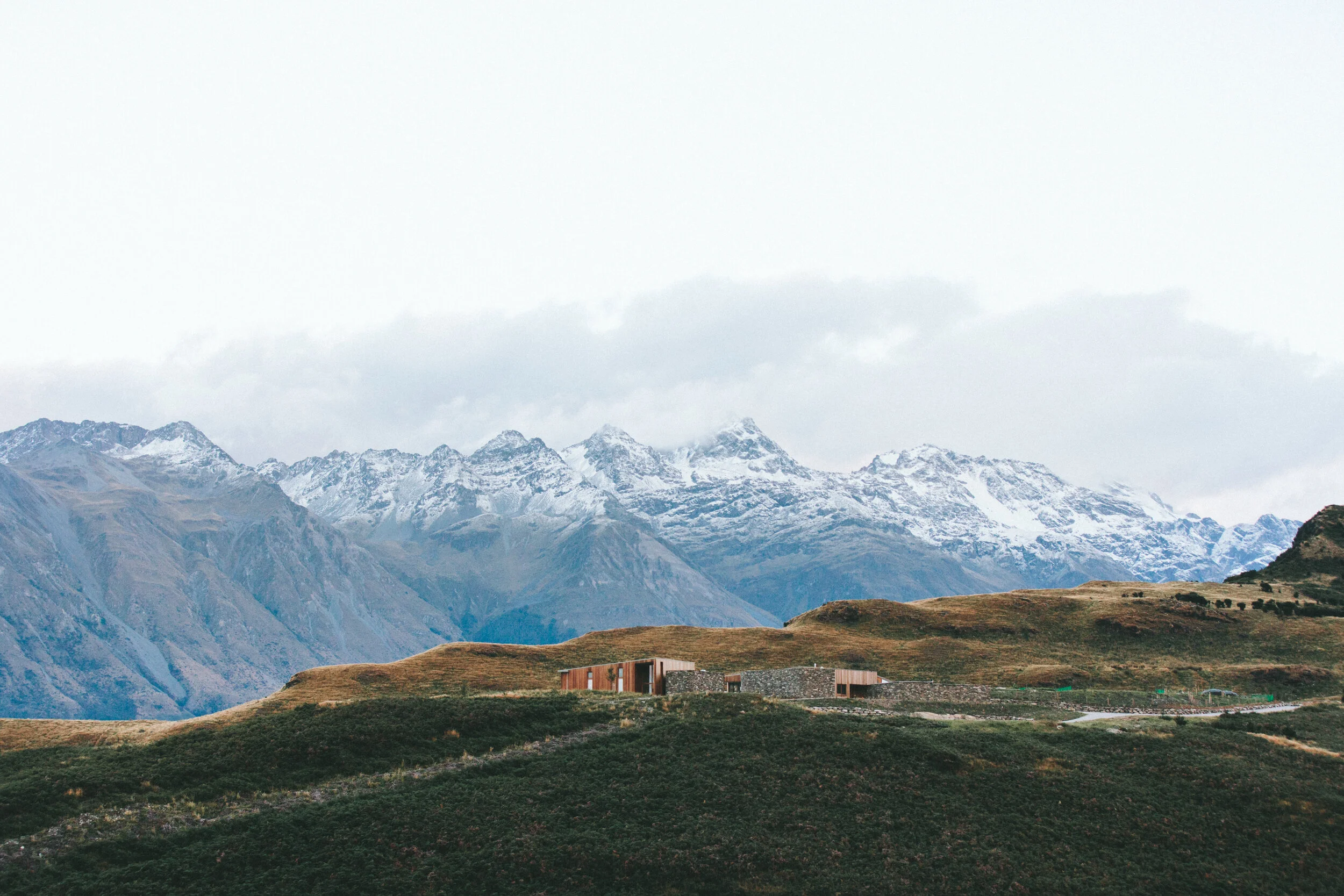 Snow-capped mountains with a modern house on a grassy hill in the foreground.
