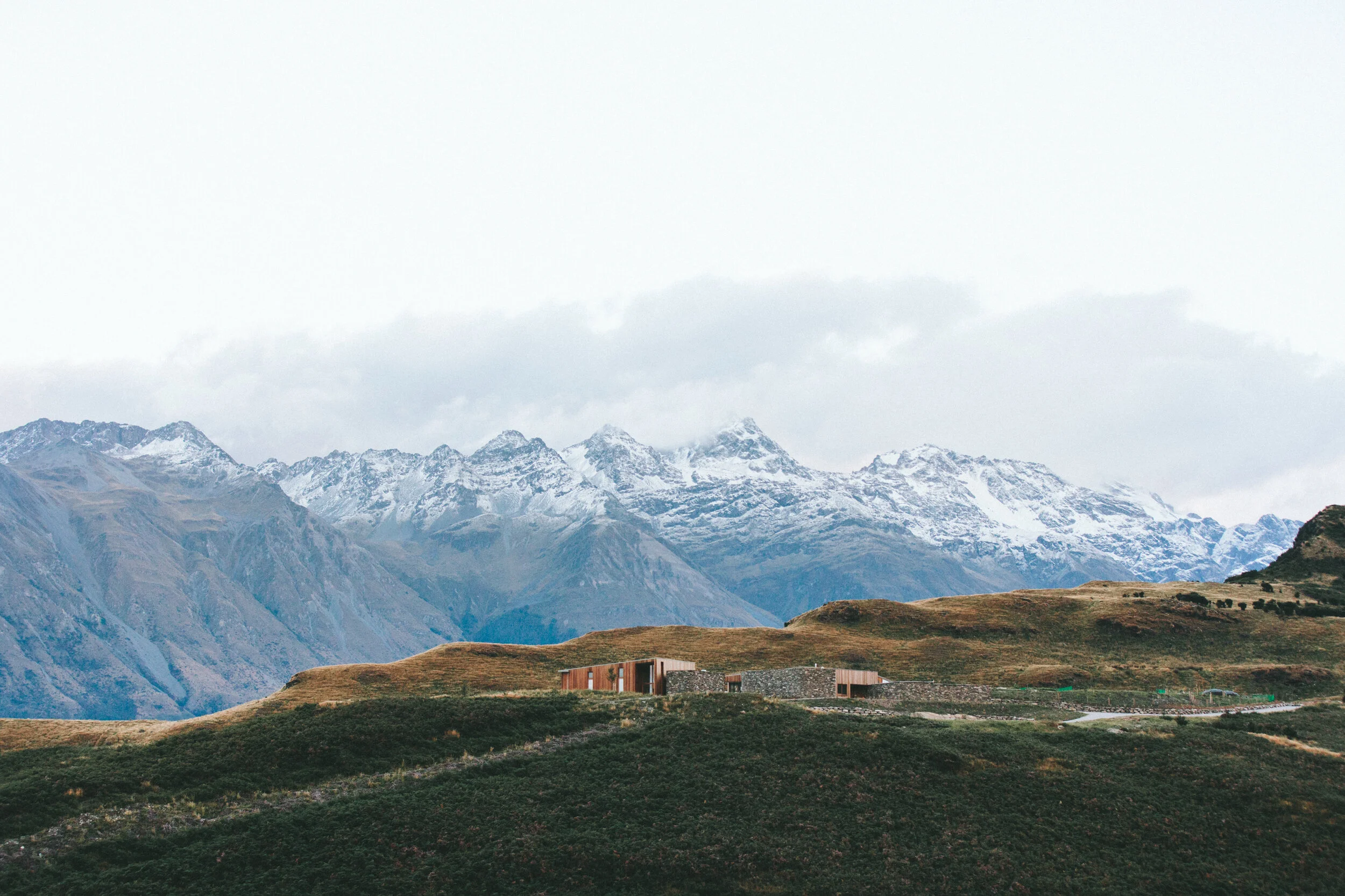 Snow-capped mountains with a modern house on a grassy hillside in the foreground.