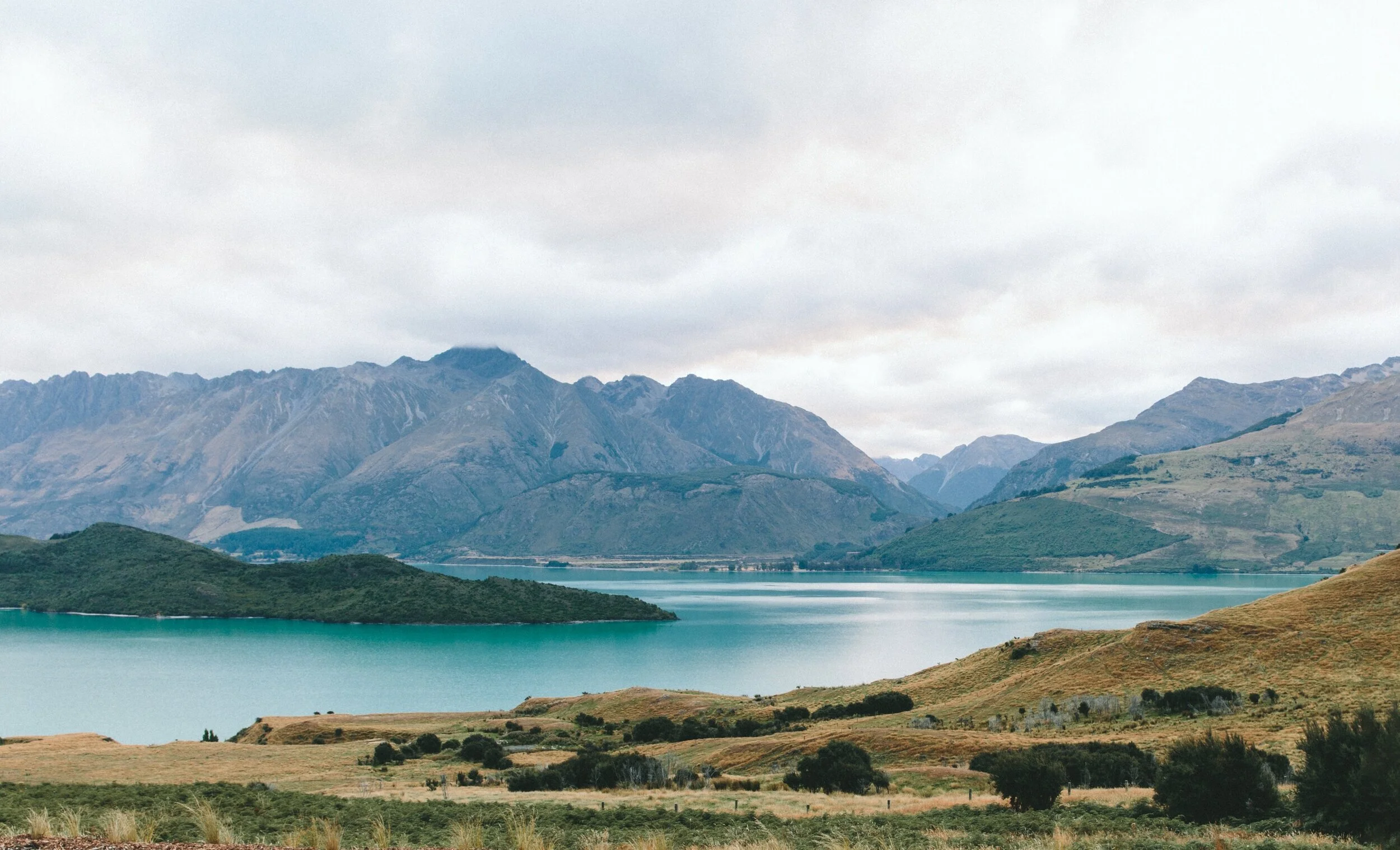 Scenic landscape with teal lake and mountain range under cloudy sky.