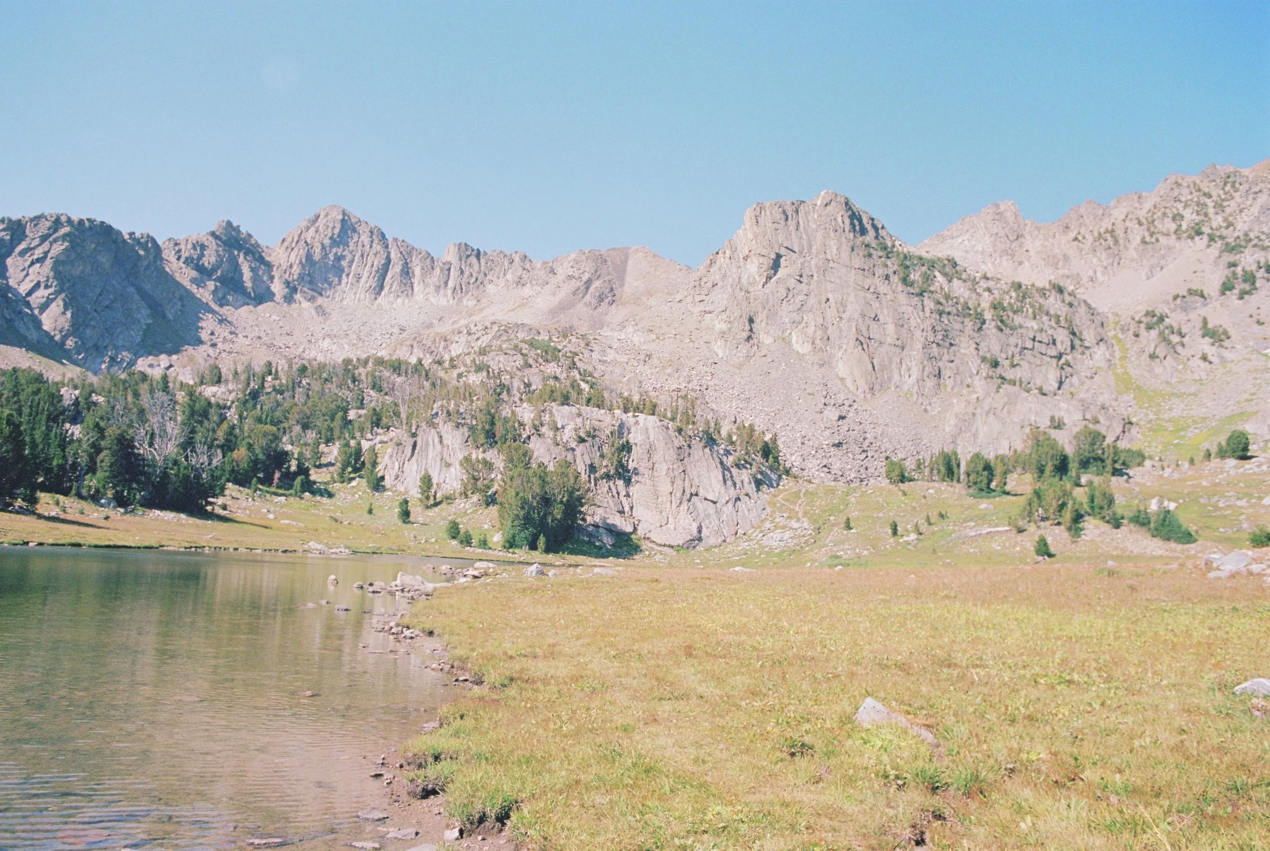 Scenic view of jagged mountain range with rocky peaks, forested hills, and a calm lake in the foreground under a clear blue sky.