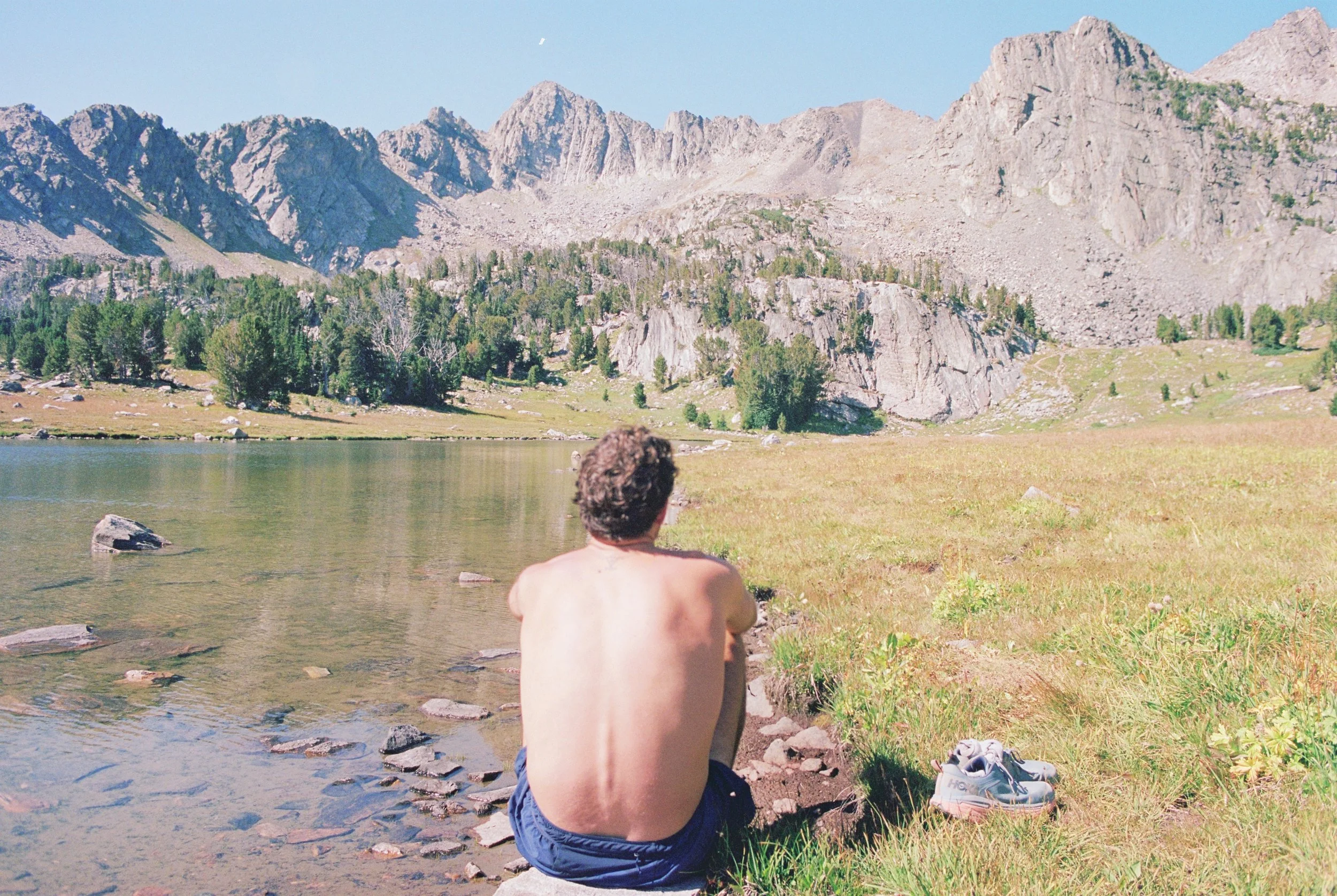 A shirtless person sits by a mountainous lake with rocky peaks in the background.