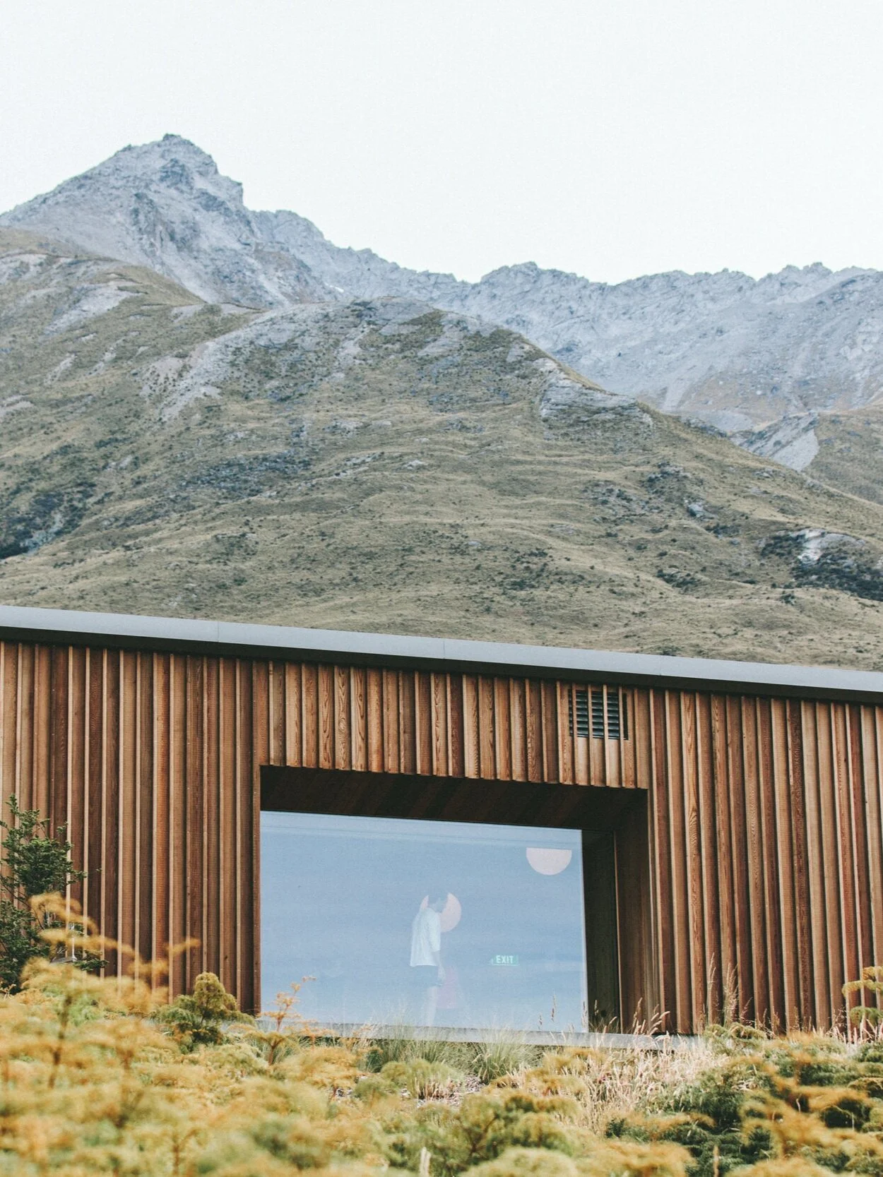 Wooden building with large window reflecting indoor scene, set against mountainous landscape
