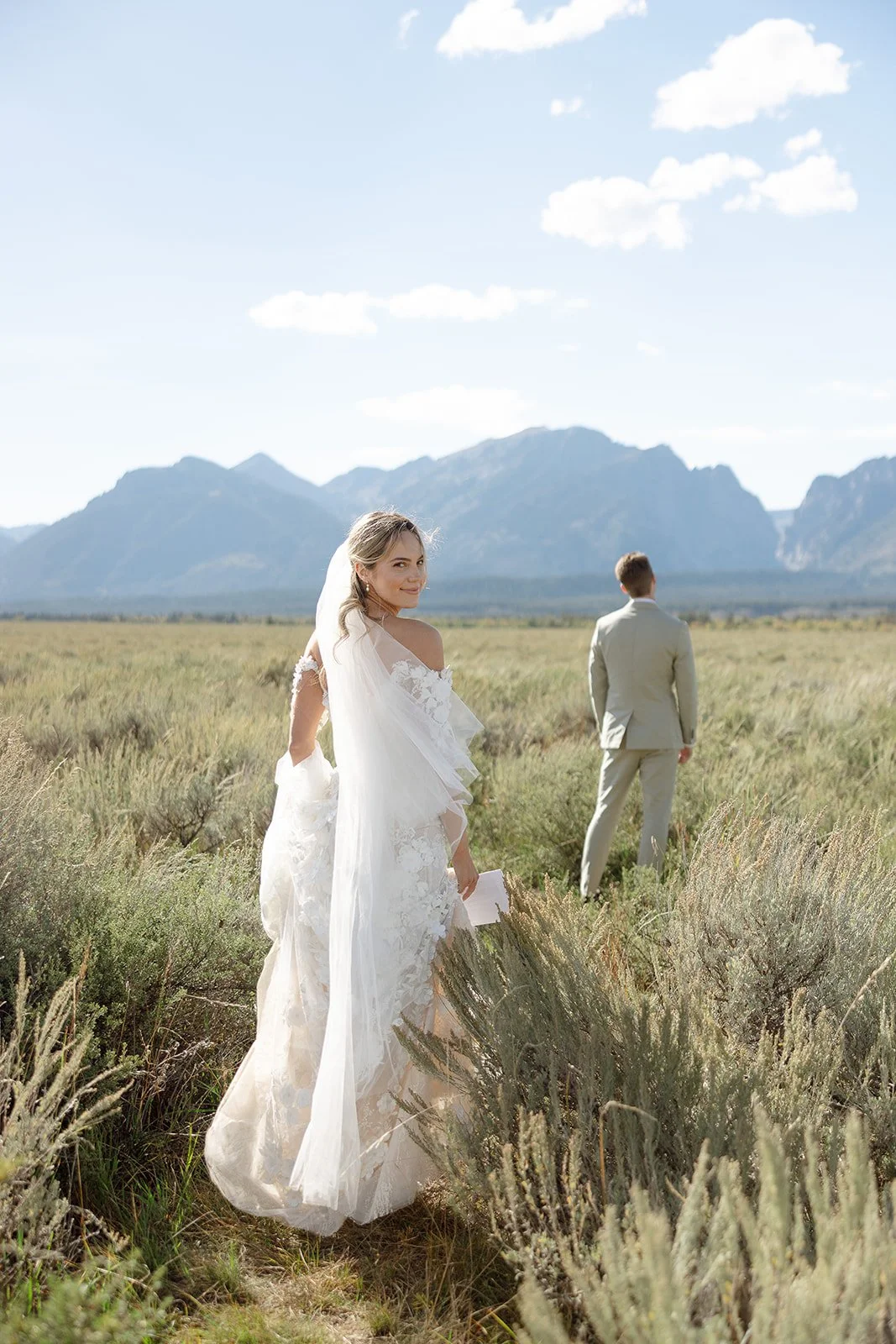 Bride and groom in a mountainous landscape, bride smiling at camera, wearing a white wedding dress with veil, groom in a light suit facing away, surrounded by grassy field and mountains in the distance.