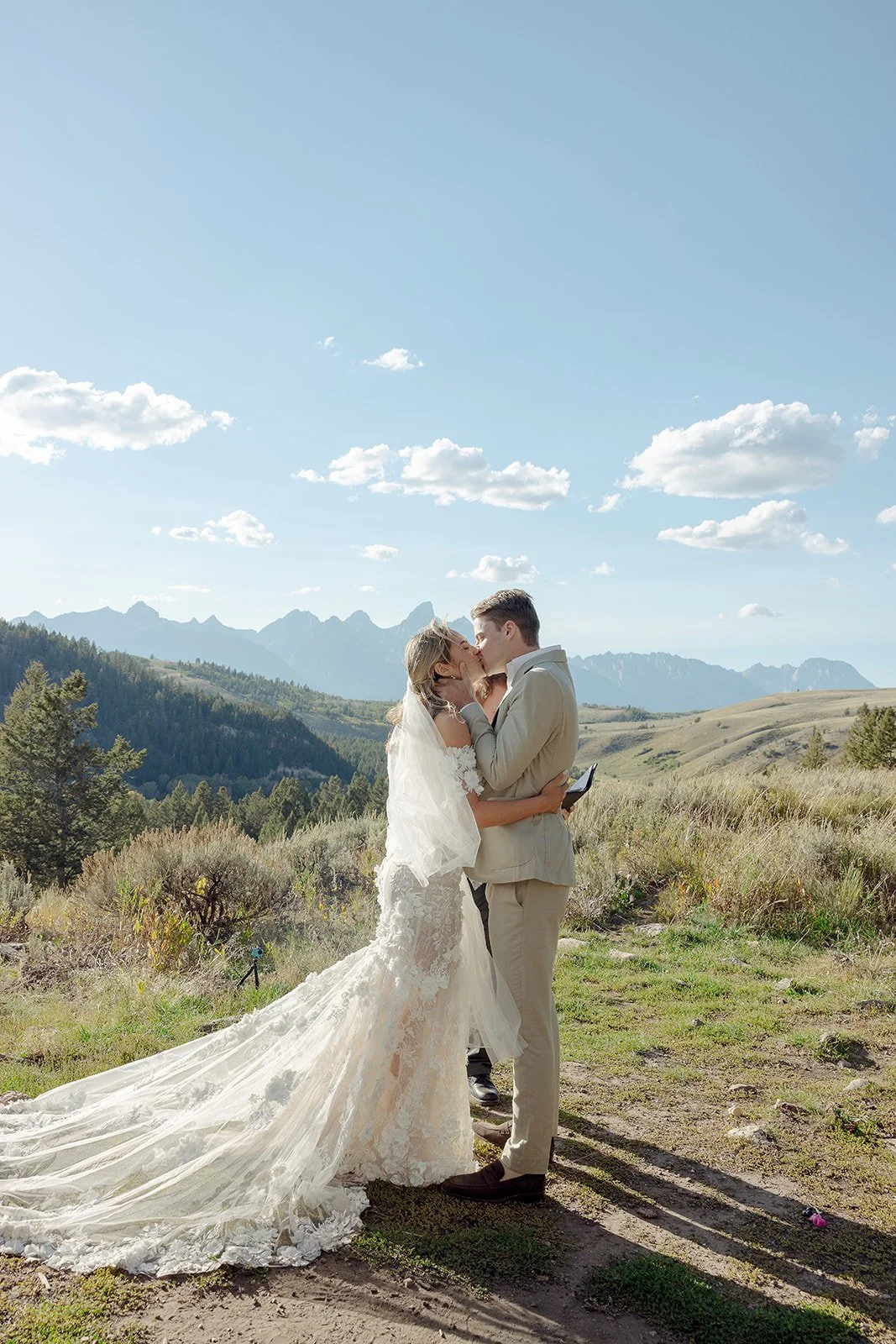 A bride and groom share a kiss in a scenic outdoor mountain setting on their wedding day, with a clear blue sky and white clouds above.