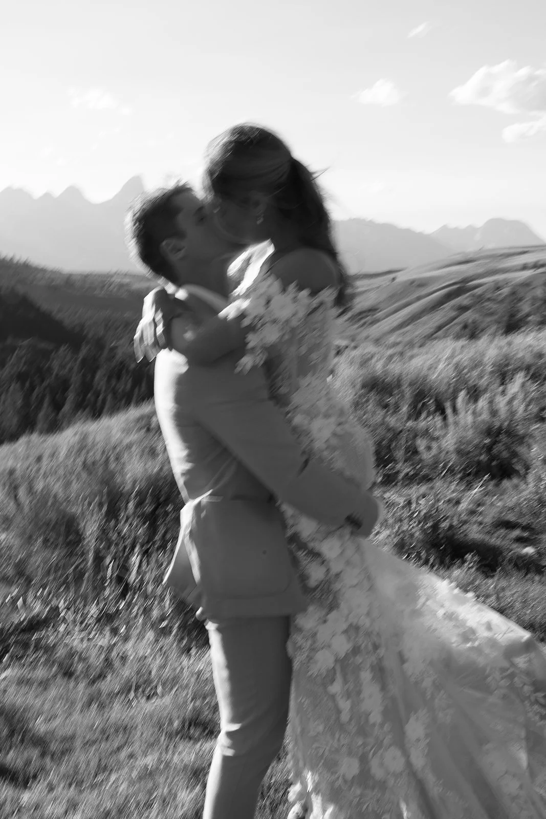 A couple, with the man lifting the woman, sharing a kiss outdoors with a scenic mountain and open field background in black and white.