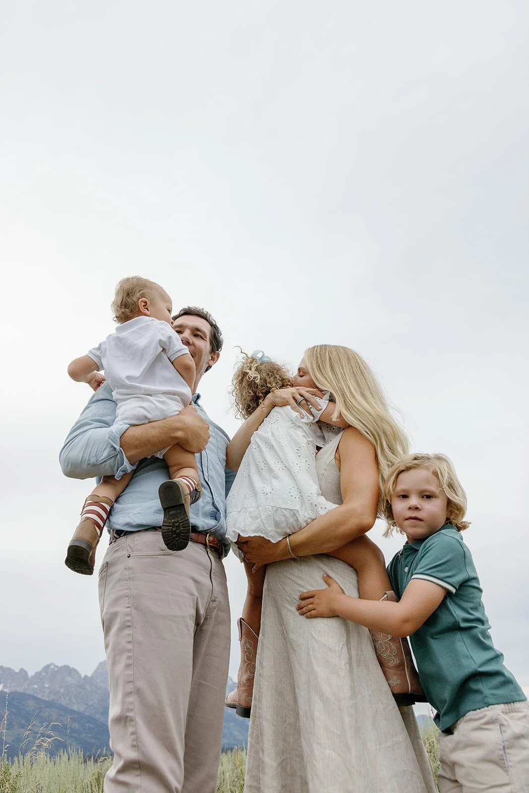 A family of five standing outdoors, sharing hugs and kisses, with mountain range in the background, under an overcast sky.