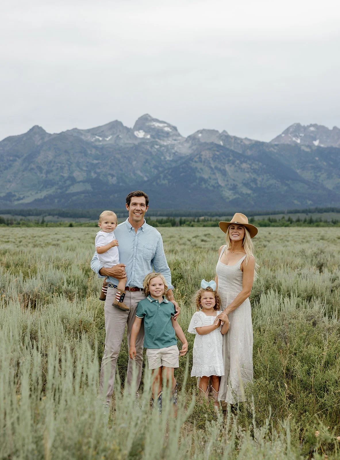 Family of five standing in a grassy field with mountains in the background, smiling at the camera.
