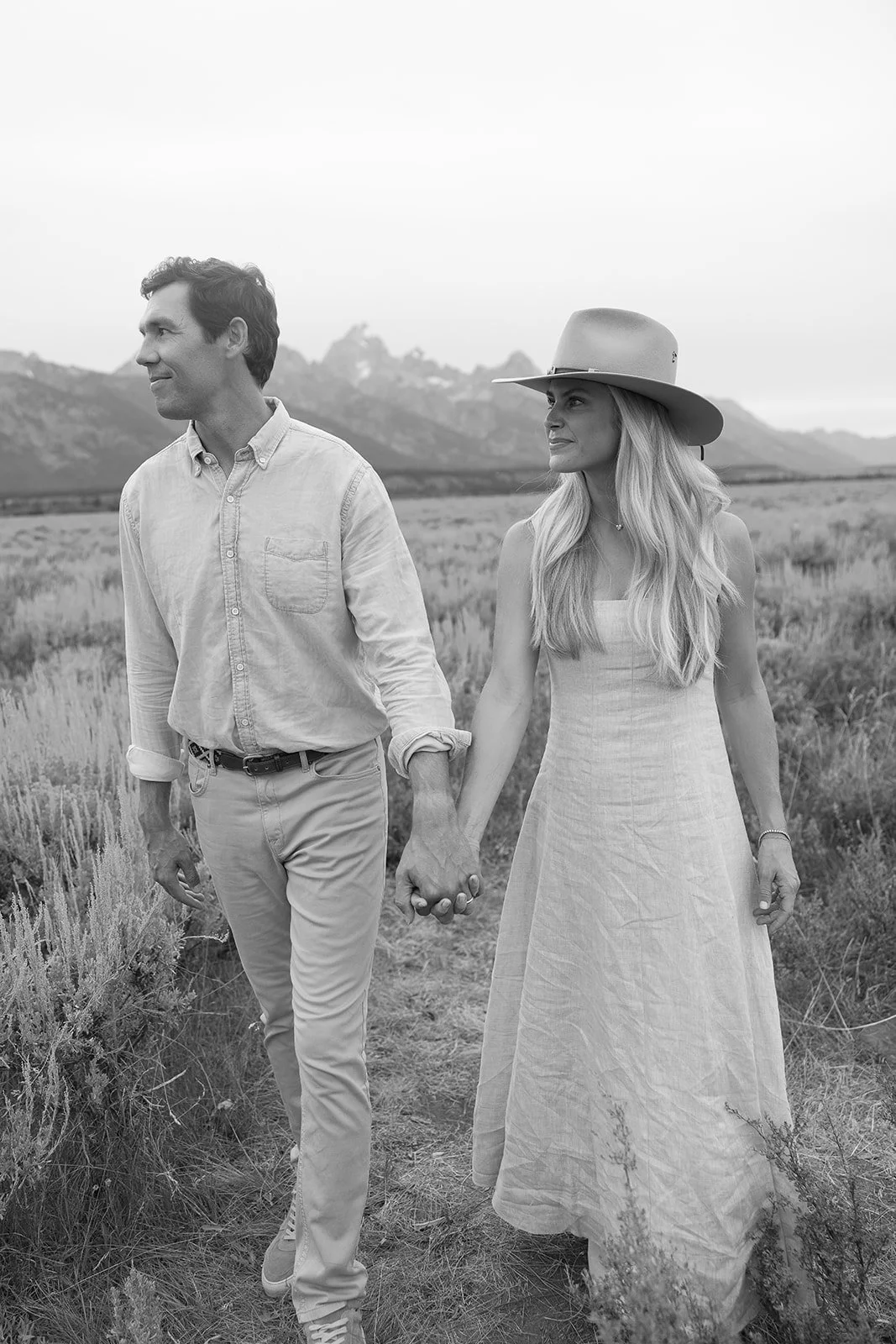 A couple holding hands and walking outdoors in a field with mountains in the background, in black and white.
