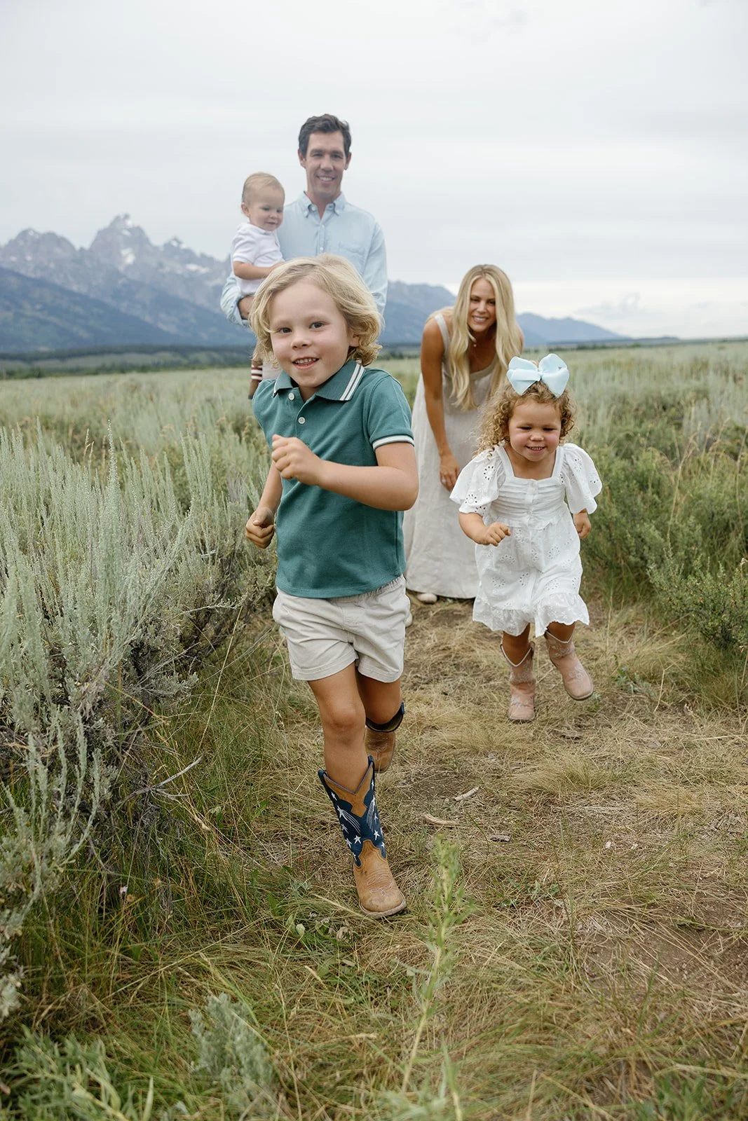 A family of five running outdoors in a grassy field with mountains in the background. The children are leading, with two girls wearing white dresses and a boy in a teal shirt and shorts, and the parents are smiling behind them, a man holding a toddle
