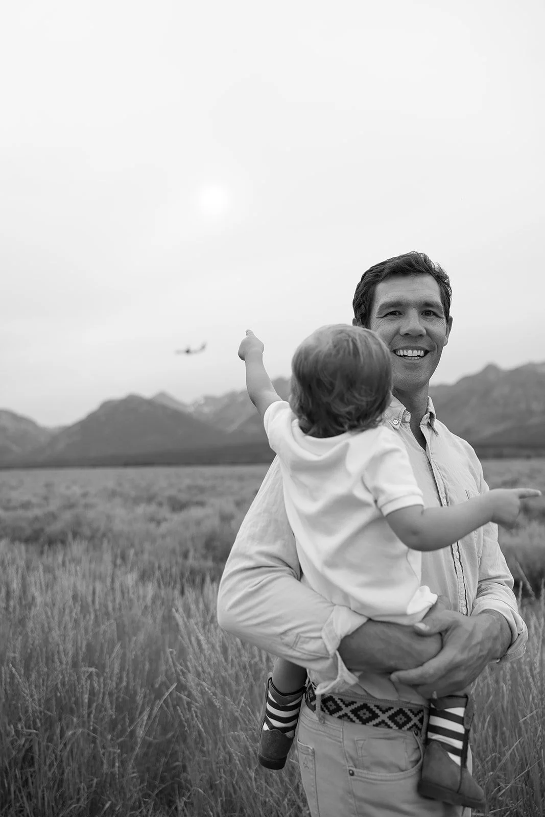 A man holding a child in a grassy field with mountains in the background. An airplane is flying in the sky.
