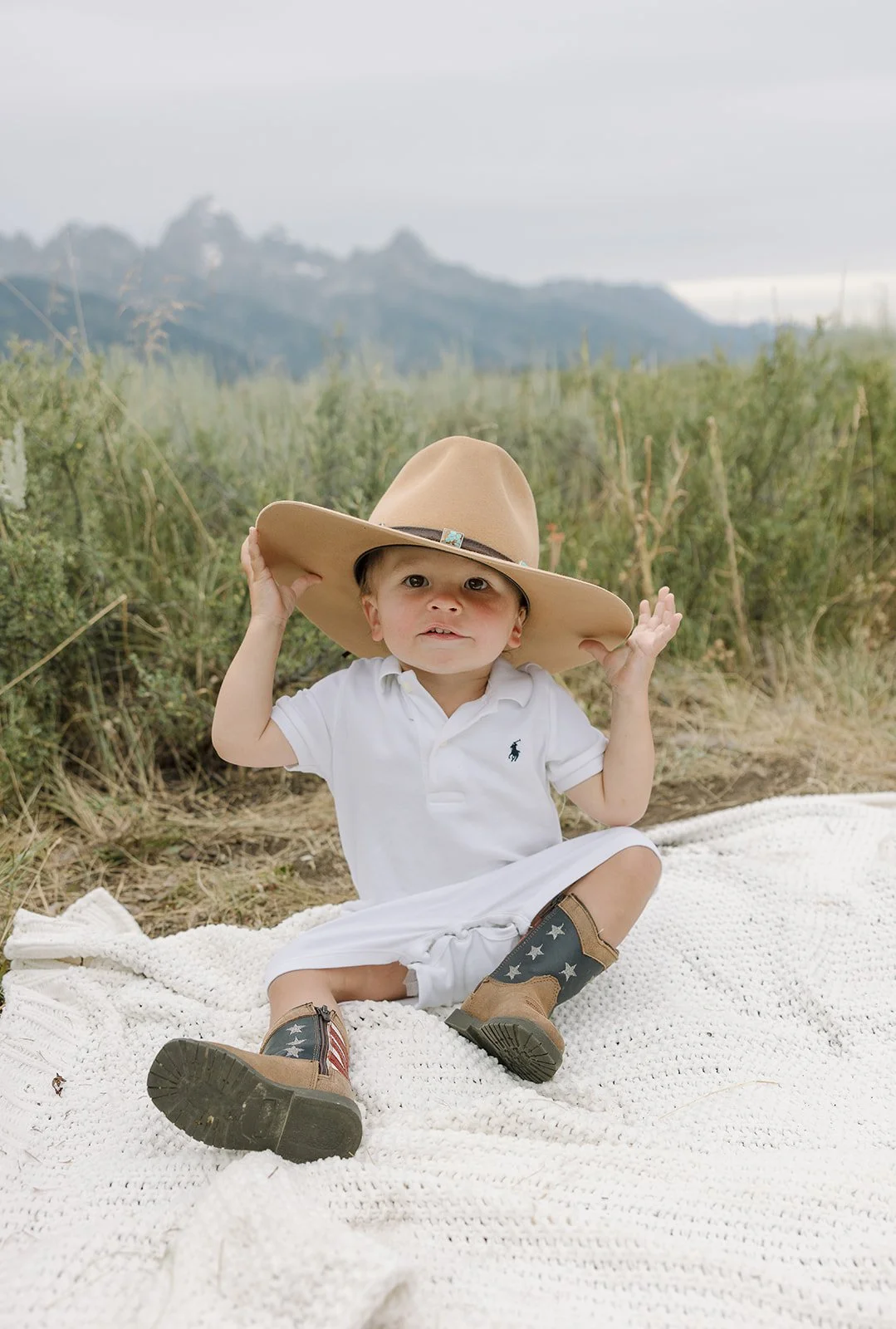 A young child sitting on a white blanket outdoors, wearing a large tan cowboy hat, a white polo shirt, khaki boots with patriotic stars and stripes, and holding the brim of the hat, with green mountains and cloudy sky in the background.