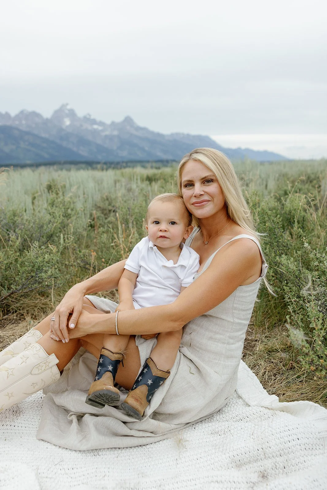 A woman and a young boy sitting on a blanket outdoors in a grassy field with mountains in the background.