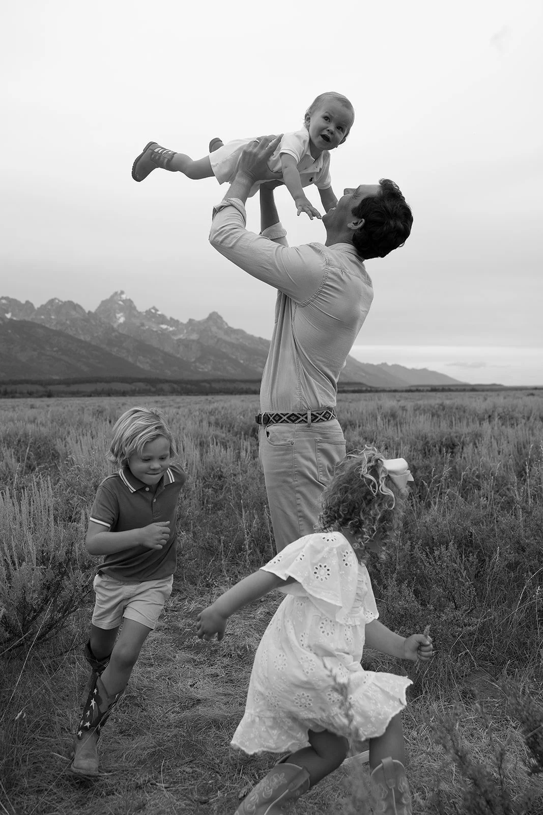 A man lifting a young child in the air outdoors with mountains in the background, and two other children running nearby.