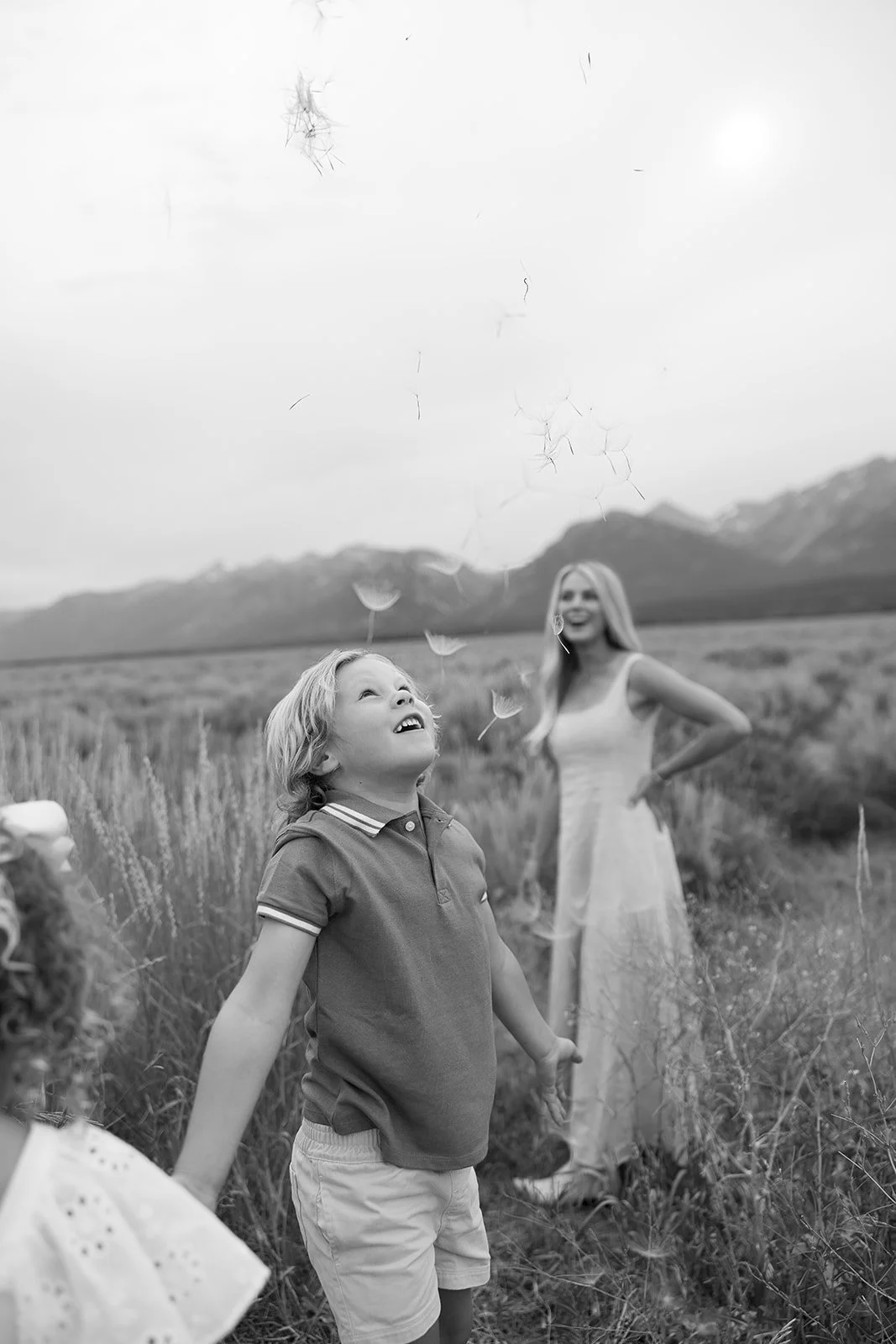 A young boy releases dandelions into the wind while a woman in a white dress looks on in an open field with mountains in the background, in black and white.