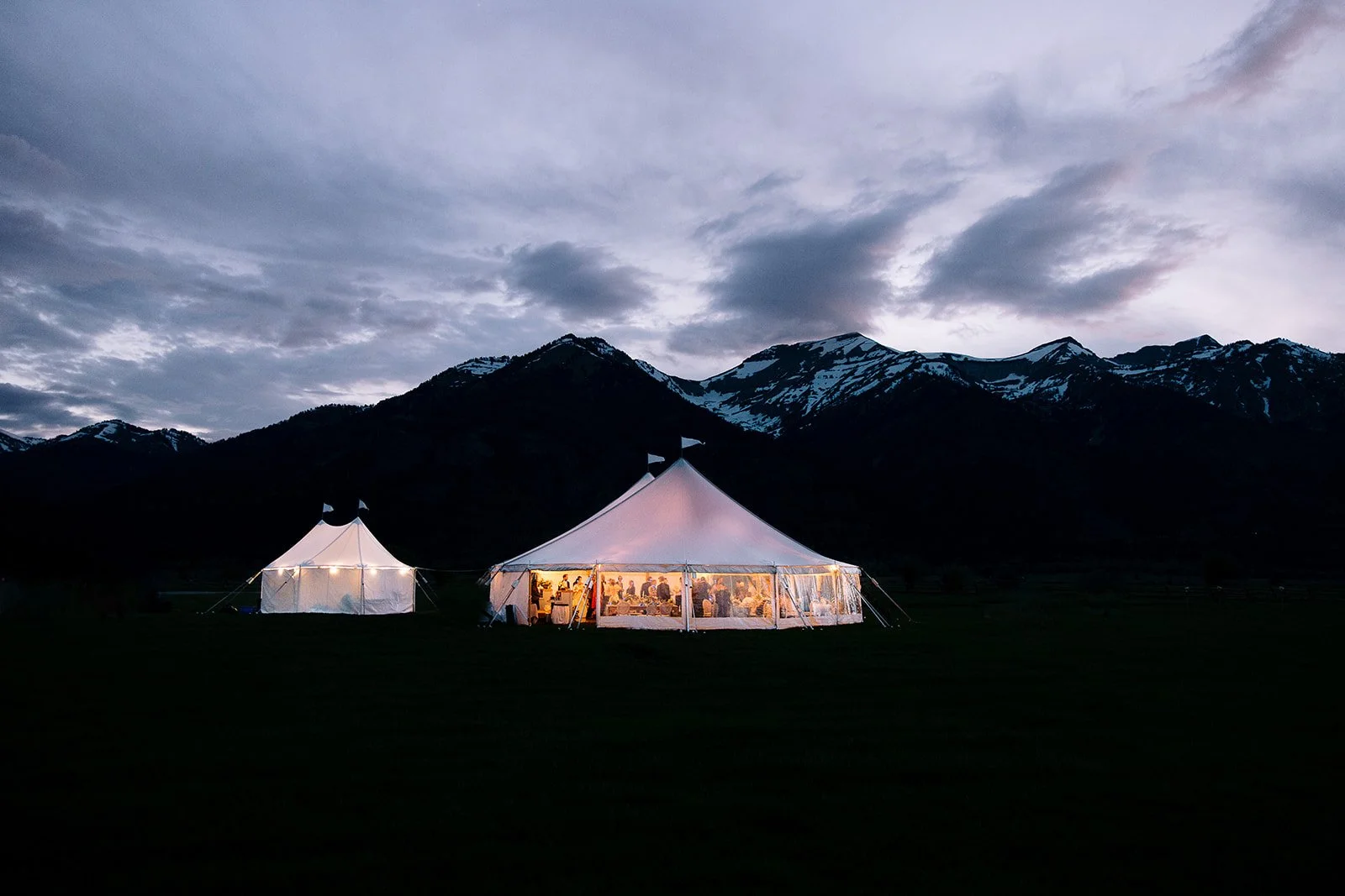 Large white event tent with transparent sides illuminated from inside, set up on a grassy field against a backdrop of mountains with snow on the peaks, under a cloudy evening sky.