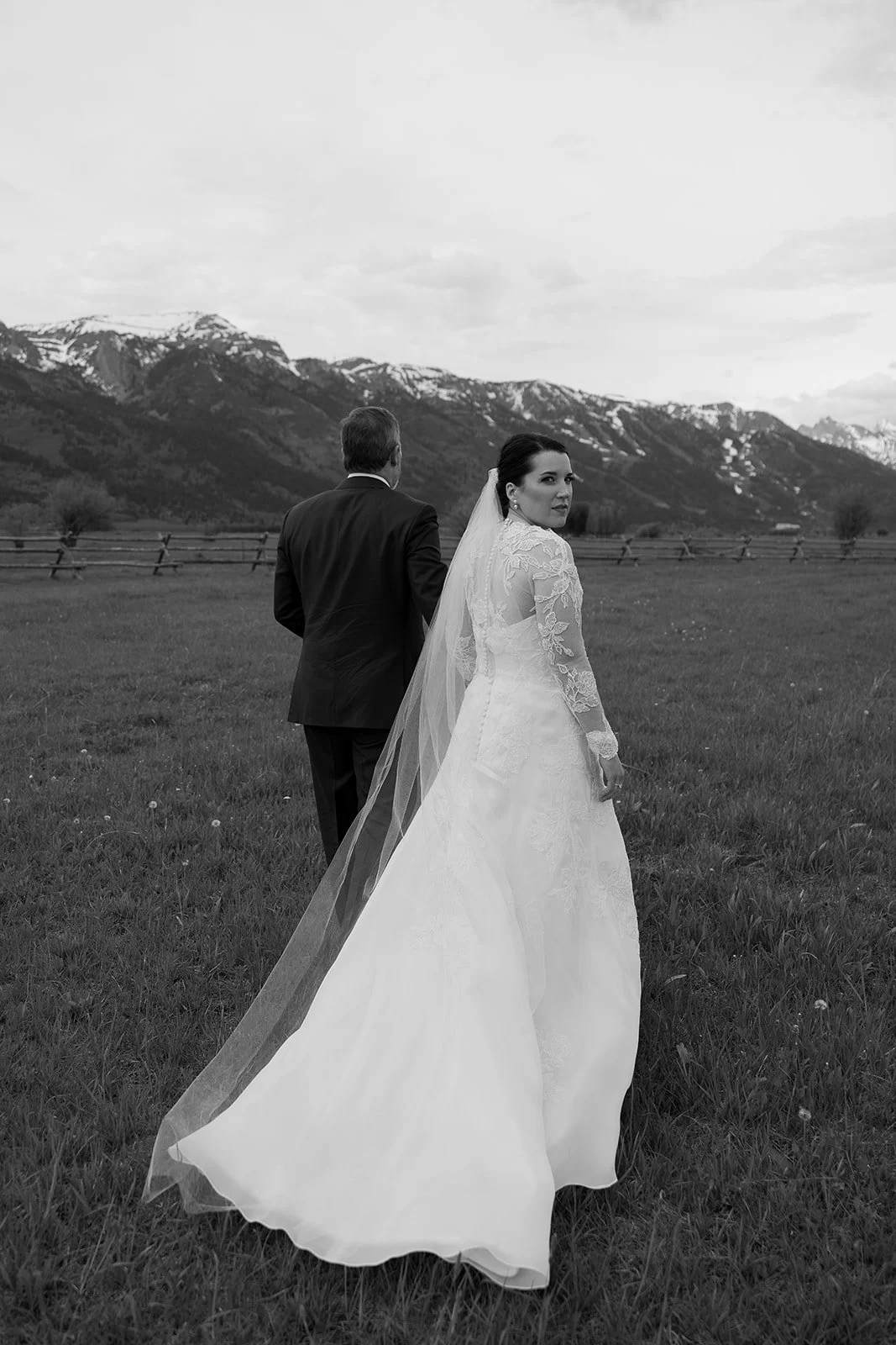 A bride and groom holding hands in an open field with mountains in the background, black and white photo.