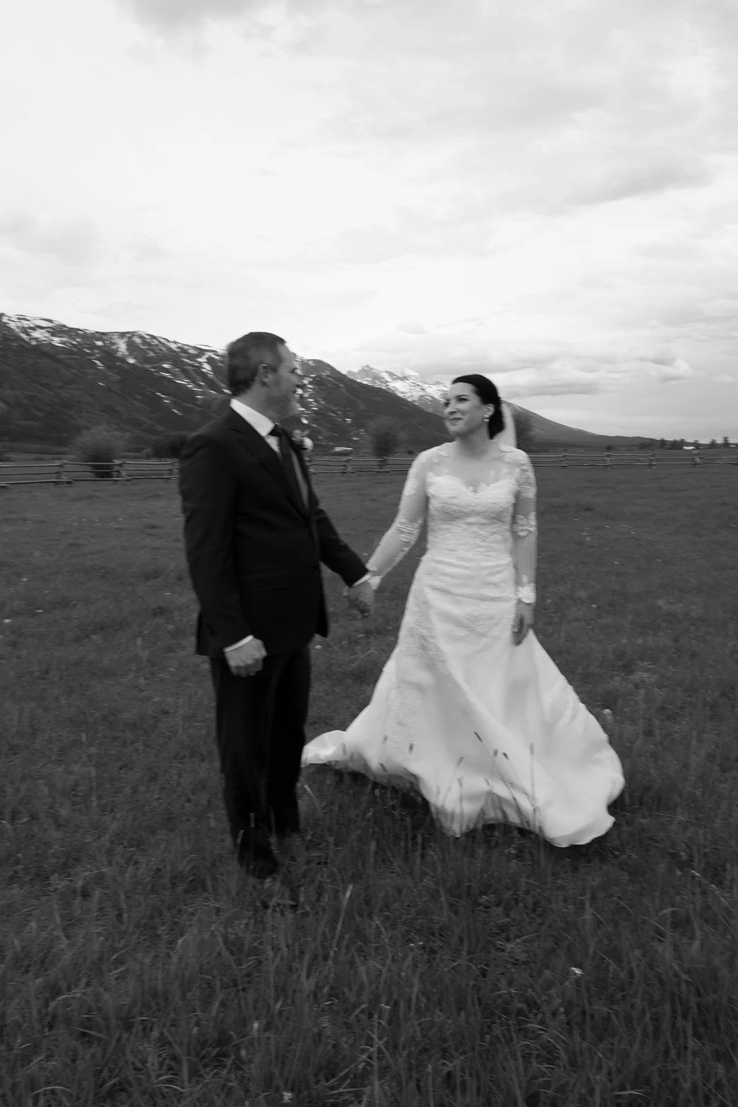 A bride and groom holding hands and looking at each other in a field with mountains in the background. The photo is in black and white.