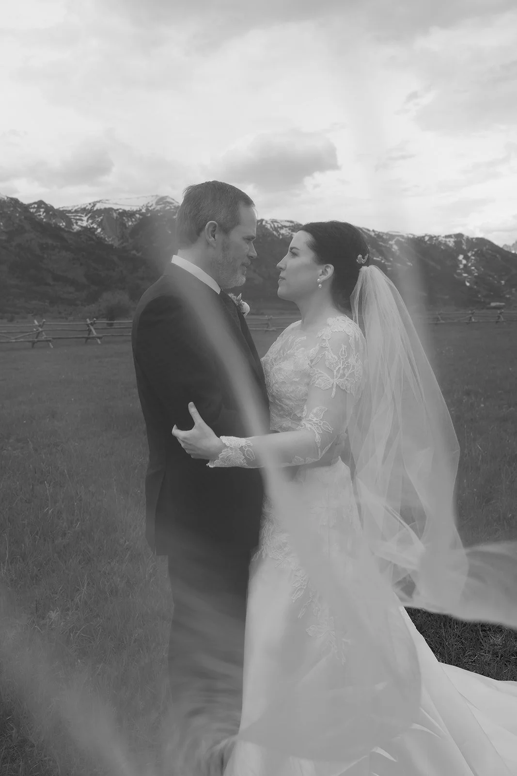 A black-and-white photo of a bride and groom standing face to face outdoors with mountains in the background. The bride wears a lace wedding gown and a veil, while the groom is in a suit. They are gazing at each other lovingly, holding each other.
