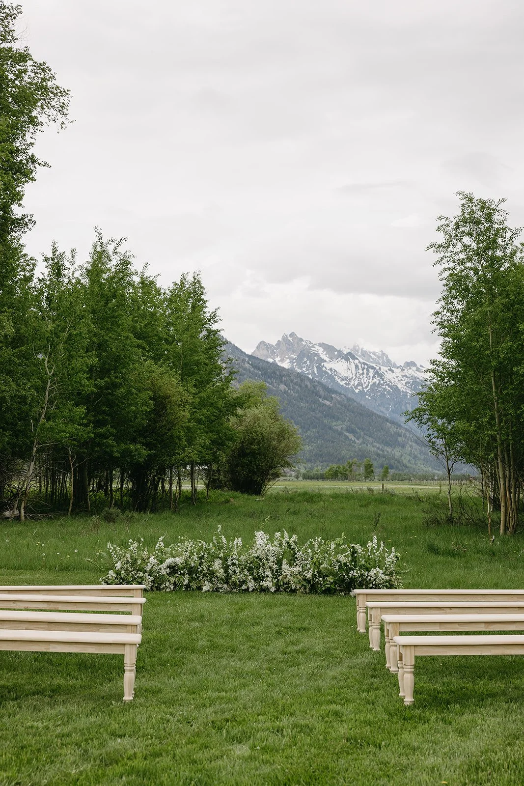 Outdoor wedding ceremony setup with white benches and floral arrangements on a green lawn, surrounded by trees with mountains in the background and overcast sky.