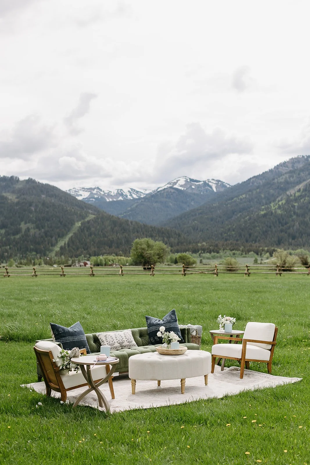 Outdoor living room setup on a grassy field with a mountain range and cloudy sky in the background. Contains a green tufted sofa with blue and white pillows, a white ottoman, two wooden armchairs with white cushions, and small side tables with floral