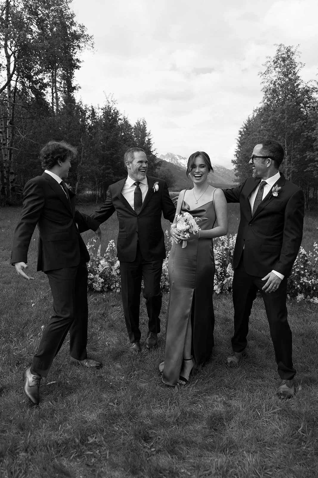 Black and white photo of a wedding party standing outdoors in a field, with trees and mountains in the background. The group includes three men in suits and a woman in a gown holding a bouquet, all smiling and holding hands, appearing to enjoy a joyf