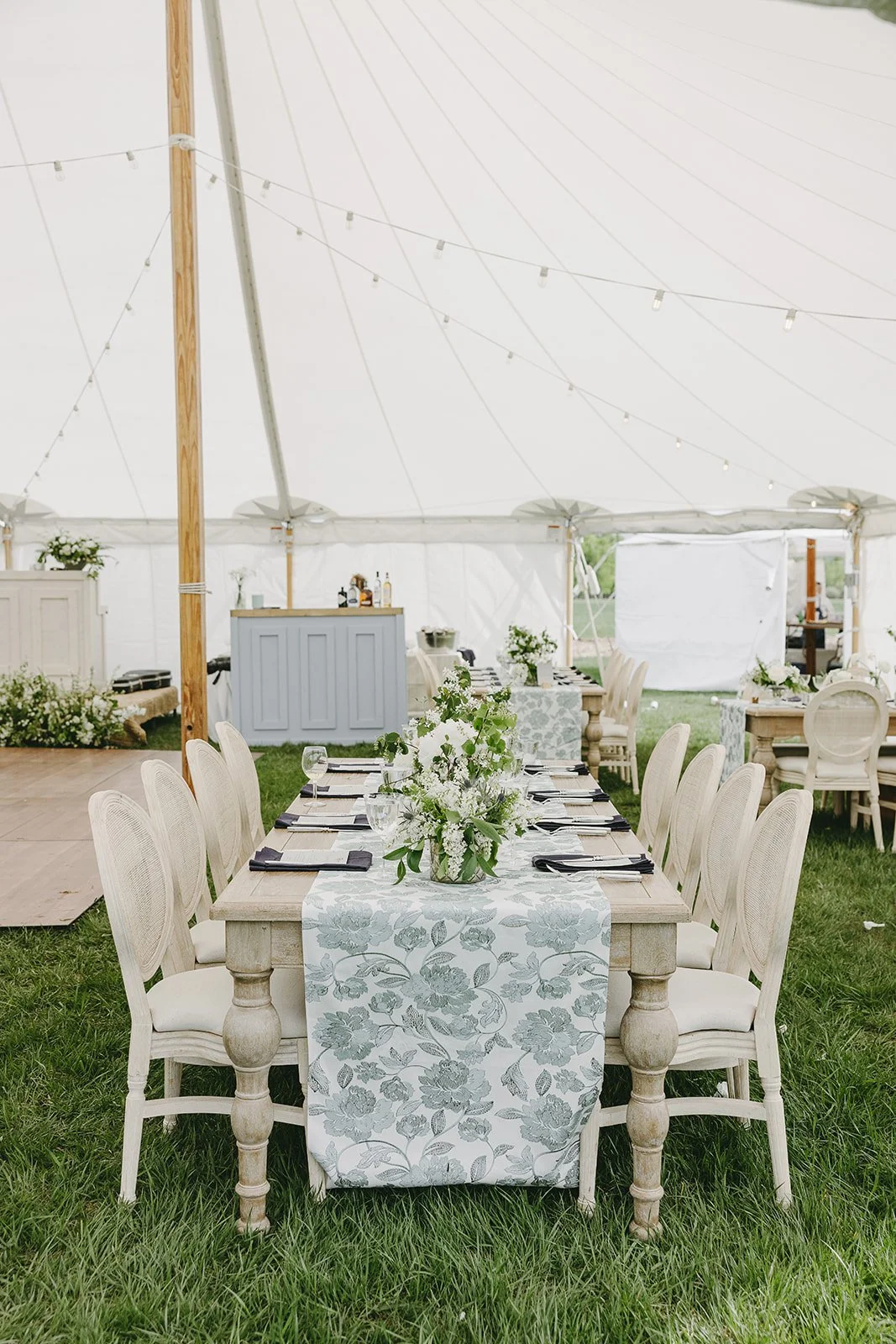 A decorated outdoor event tent with a long wooden table, white chairs, and floral centerpieces, set for a celebration.