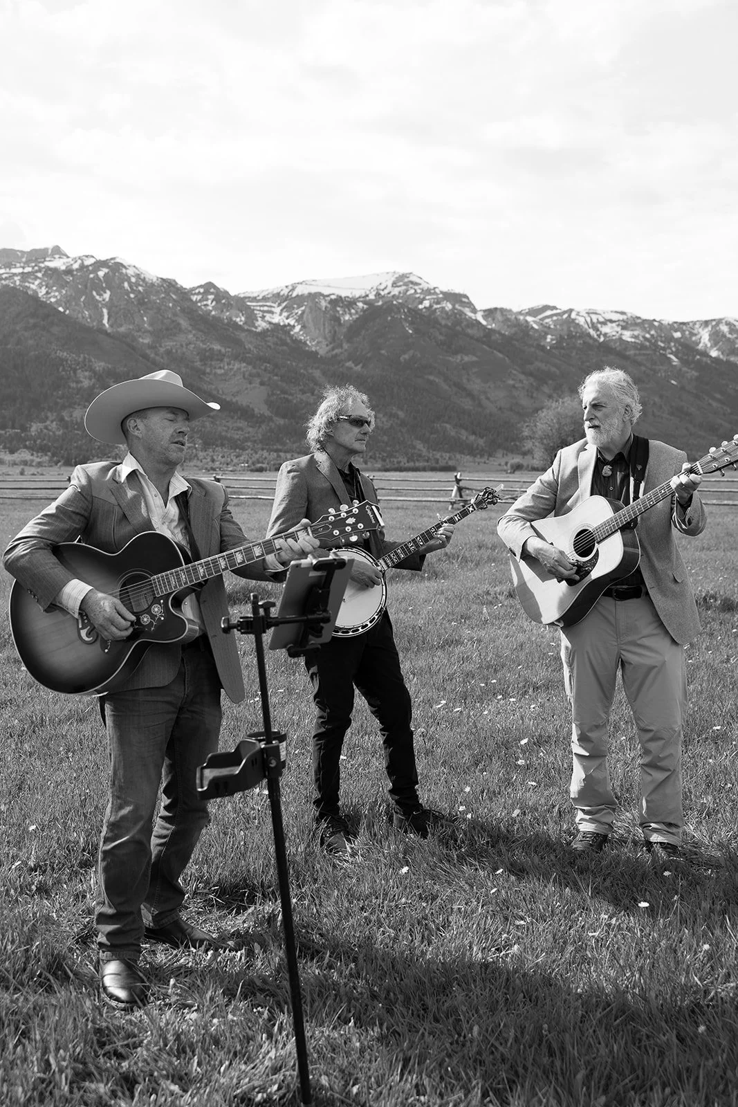 Three men playing guitars and banjo outdoors in a grassy field with mountains in the background.