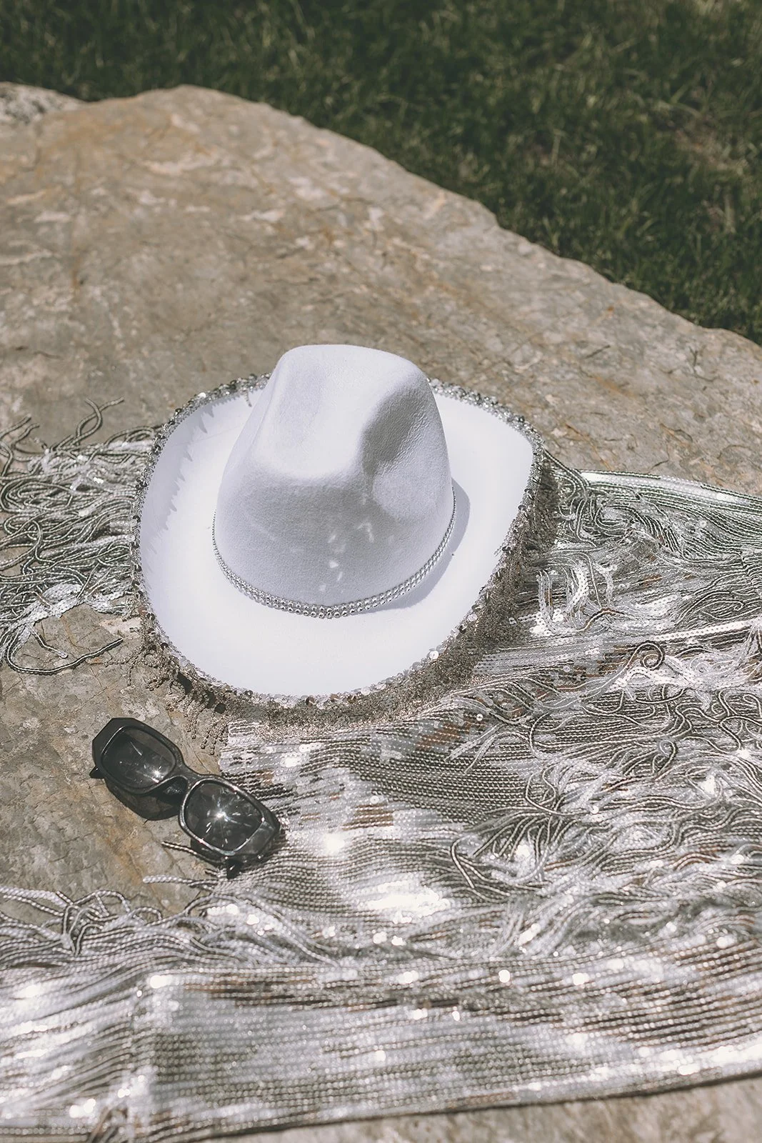 A white cowboy hat with a beaded band, silver jewelry, and black sunglasses placed on a silver fringed fabric, all resting on a stone surface outdoors.