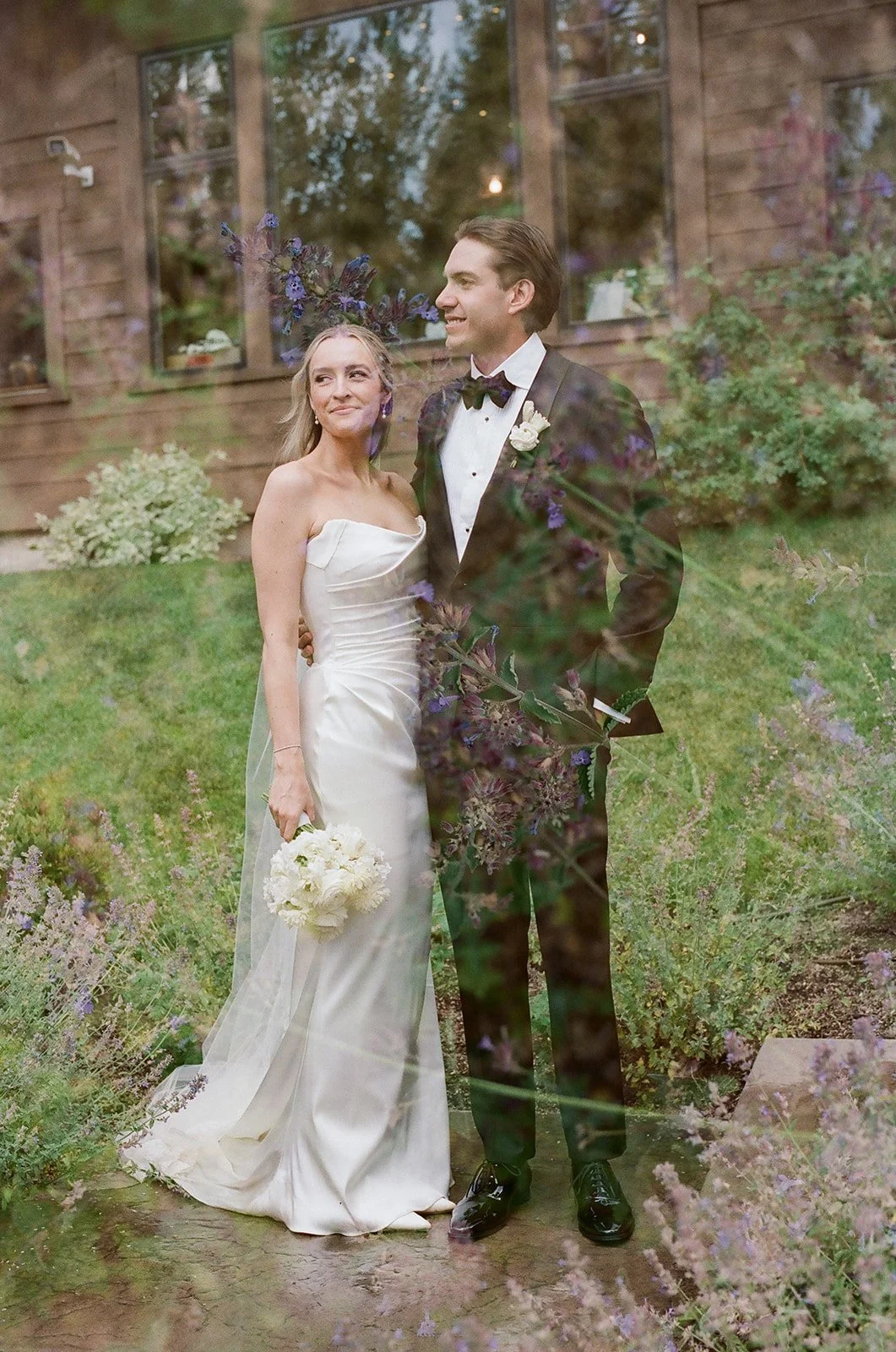 A bride and groom standing outside on a lush green lawn, behind a glass window with reflections, surrounded by purple and green foliage, during a wedding celebration.