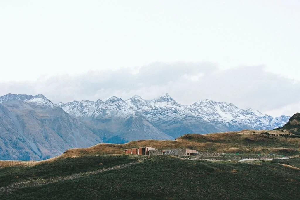 Snow-capped mountains with cloudy sky, grassy hills, and small building in foreground.