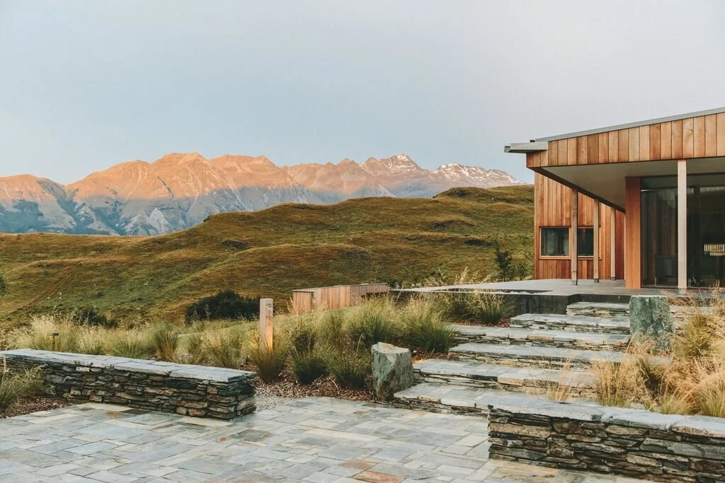 Modern wooden house with stone patio in grassy landscape, mountain view in background at sunset.