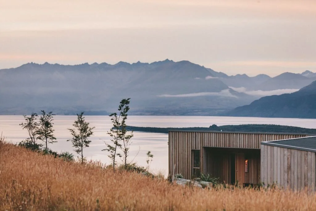 Scenic landscape of a modern wooden cabin overlooking a lake with distant mountains and a cloudy sky at sunset.