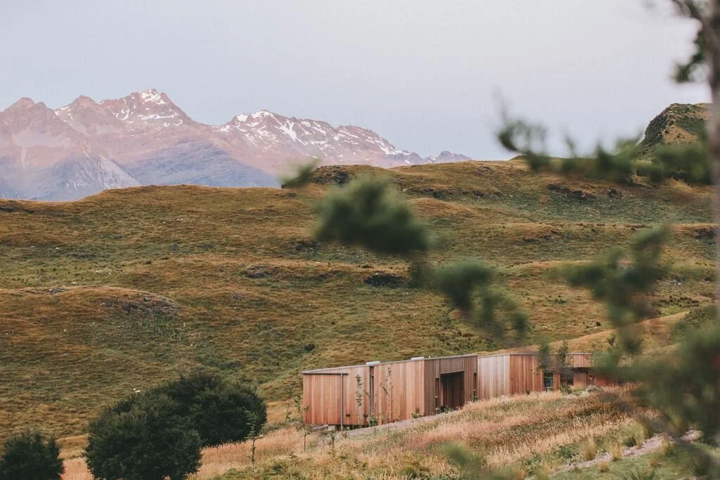 Wooden cabin amidst grassy hills with snow-capped mountains in the background.