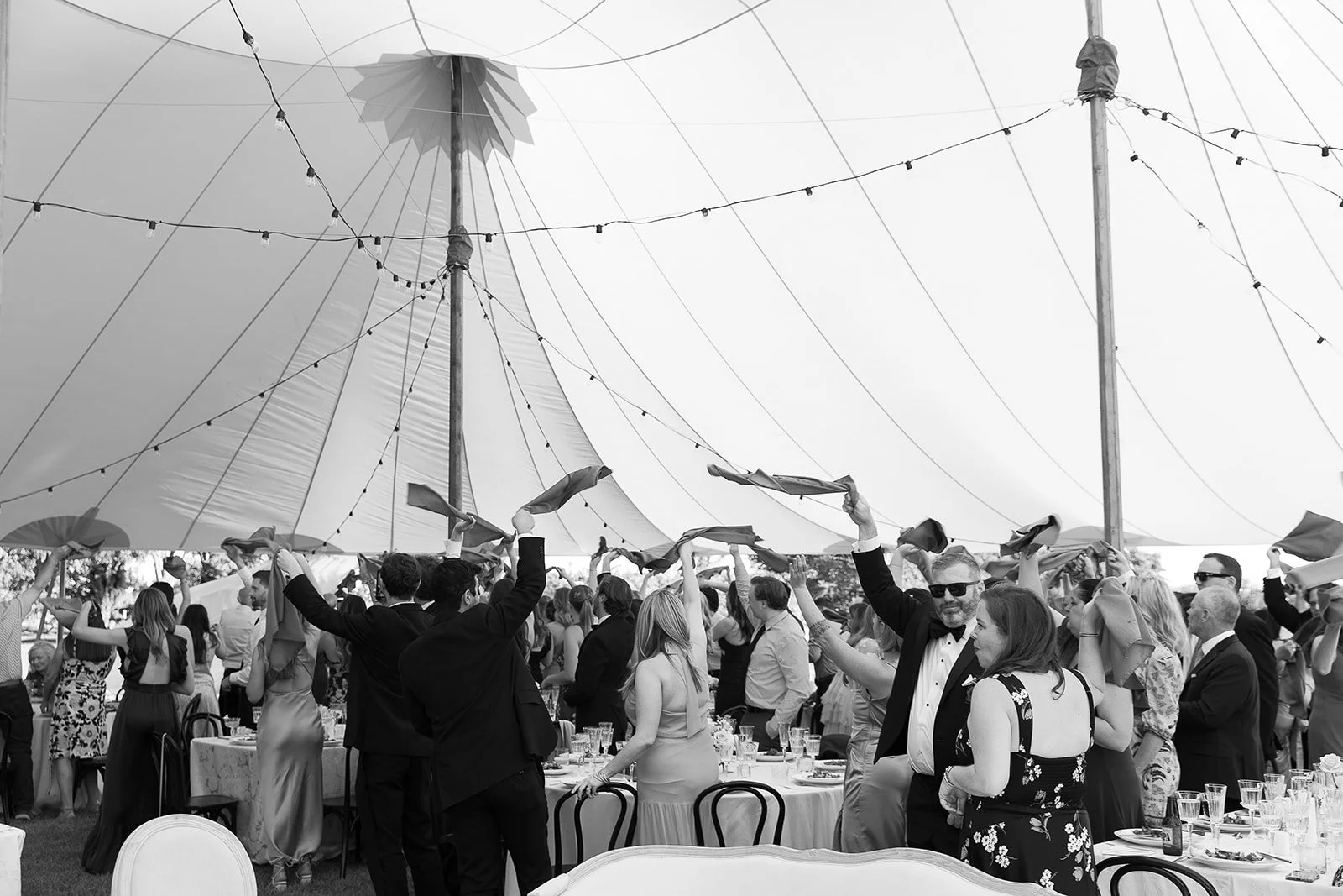 Black and white photo of a lively wedding reception under a large tent. Guests are standing around tables, waving napkins in celebration. String lights adorn the tent ceiling.