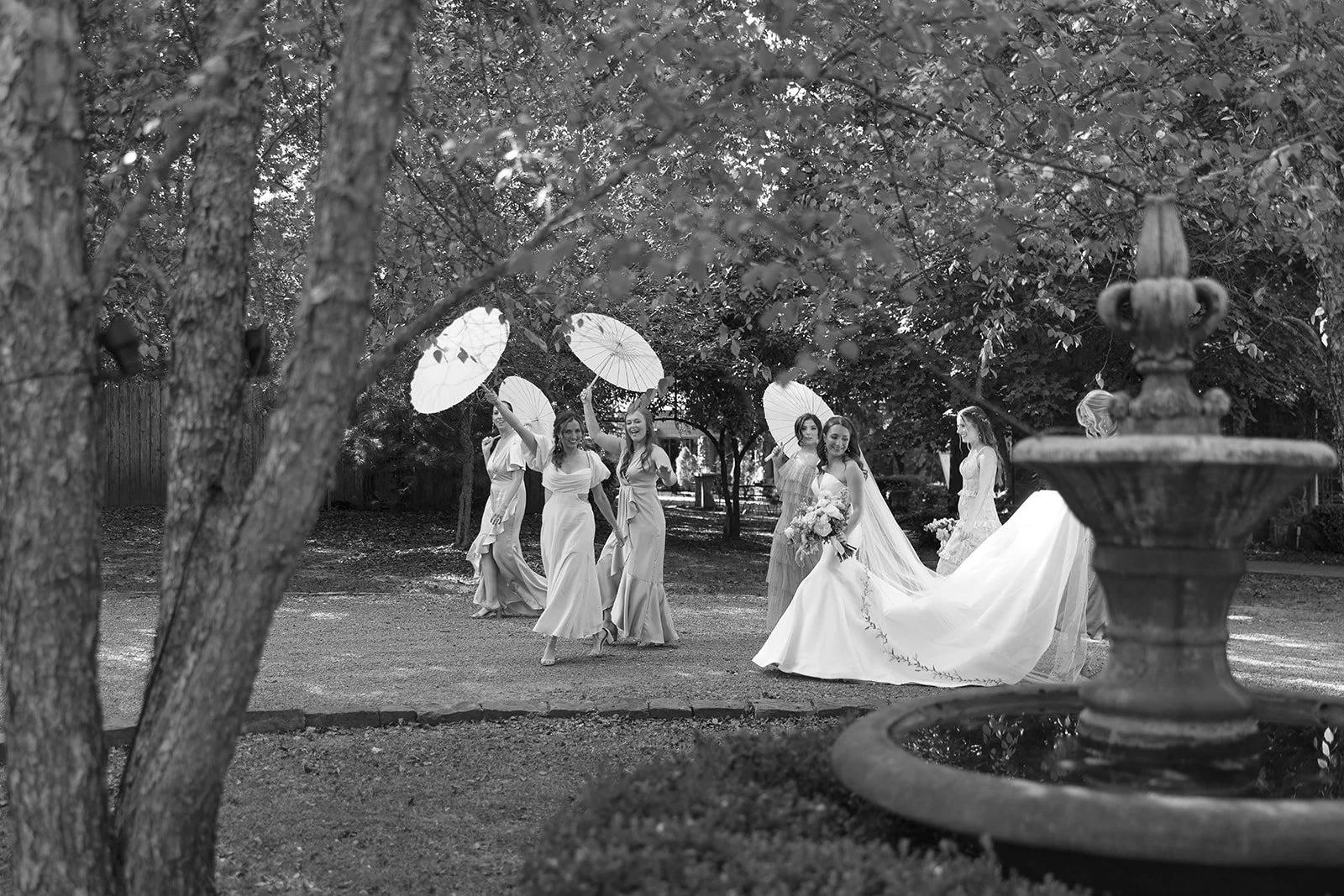 A black and white image of a bridal party in a garden. The bride is wearing a long gown and holding a bouquet, while bridesmaids walk ahead, holding parasols. Trees and a fountain are in the foreground.