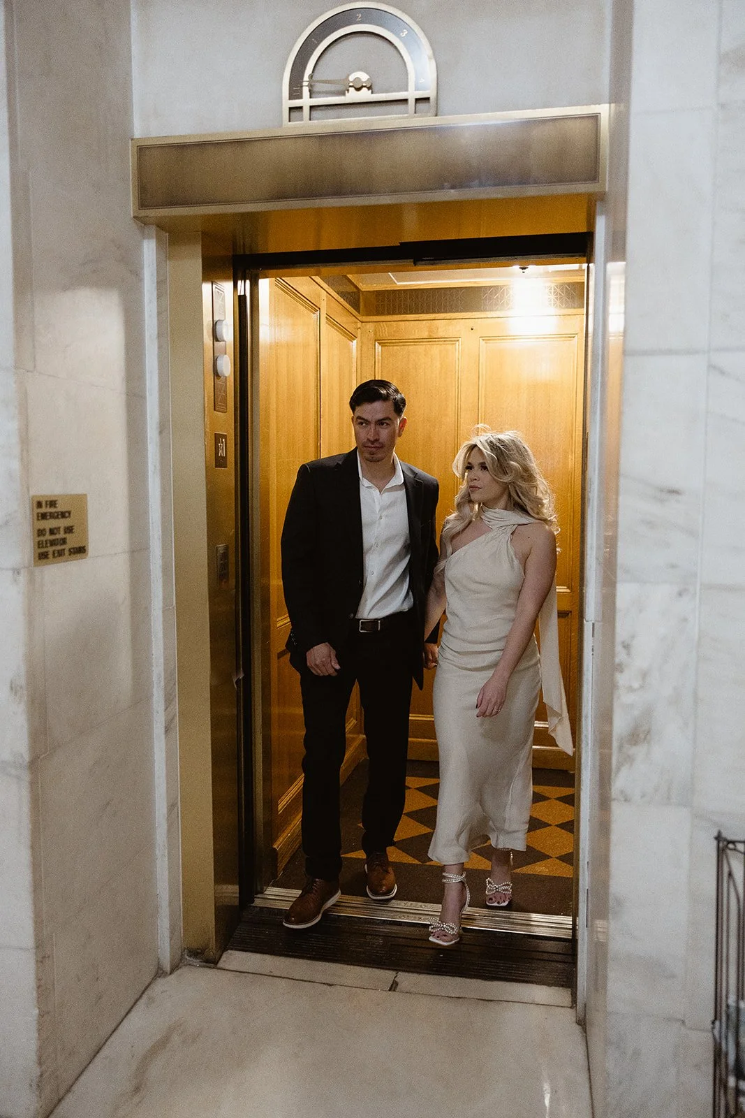 A couple exiting a wood-paneled elevator in formal attire.