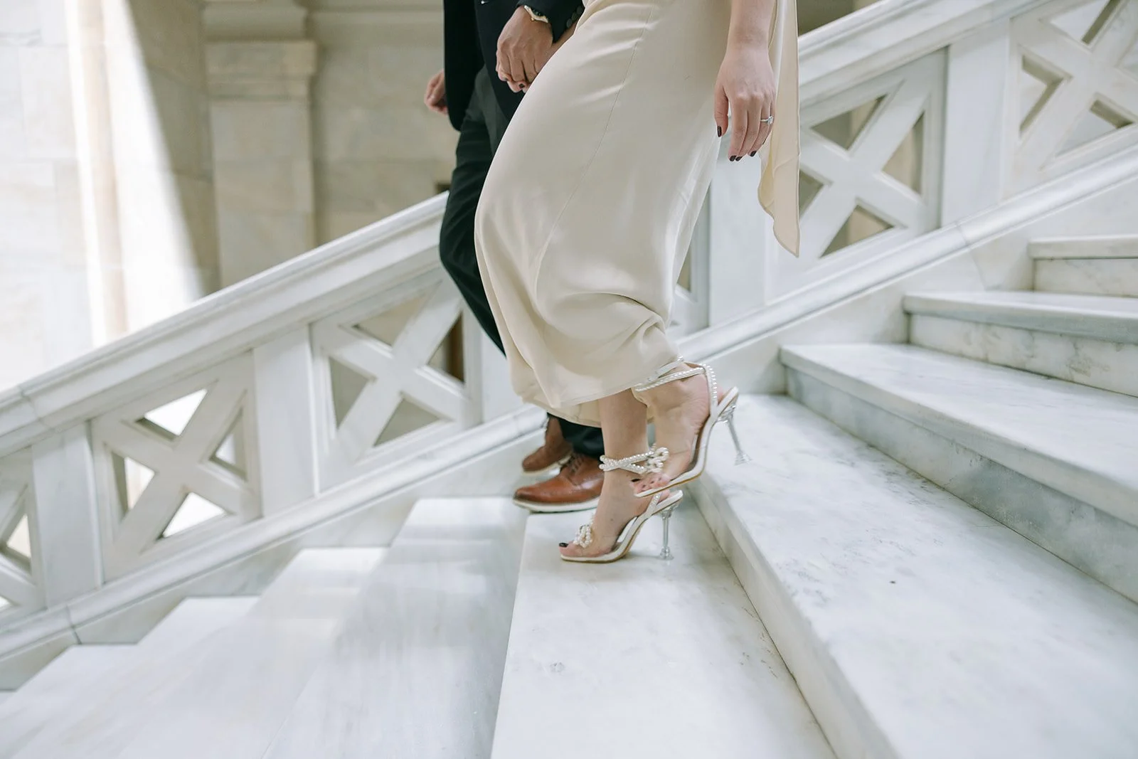 A close-up of a woman in a beige dress and high heels ascending marble stairs next to a man in black pants and brown shoes.