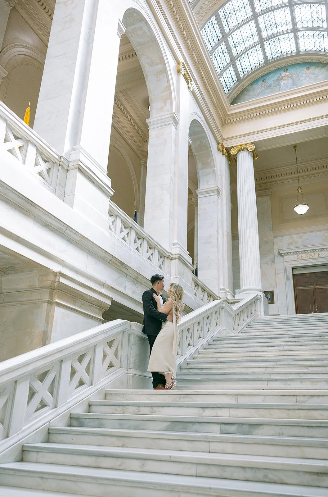 A couple embraces on a grand marble staircase inside a stately building with high arches and pillars.