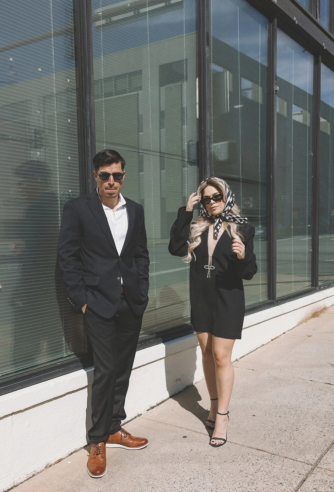 Two stylishly dressed individuals, one man and one woman, standing outside a building with large windows; the man wears a black suit and brown shoes, while the woman wears a black outfit and sunglasses with a patterned headscarf.