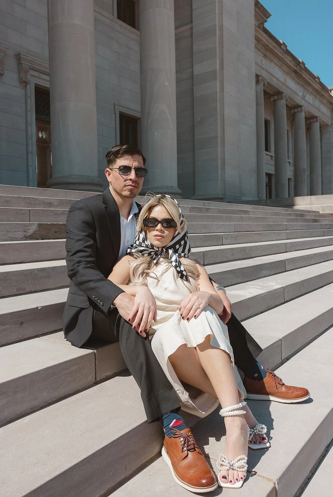 A couple sitting on steps outside a large building with columns. The man is wearing a suit and sunglasses, while the woman is in a dress, headscarf, and sunglasses.