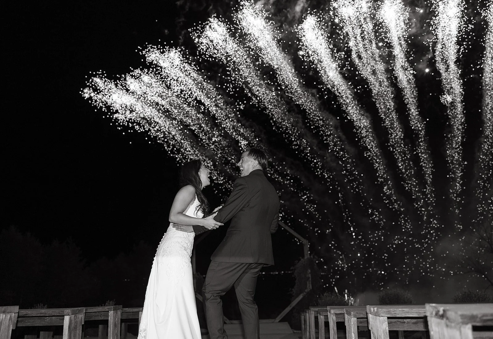 Black and white photo of a couple in formal attire, holding hands and smiling, with fireworks in the night sky overhead. The scene is energetic and celebratory.