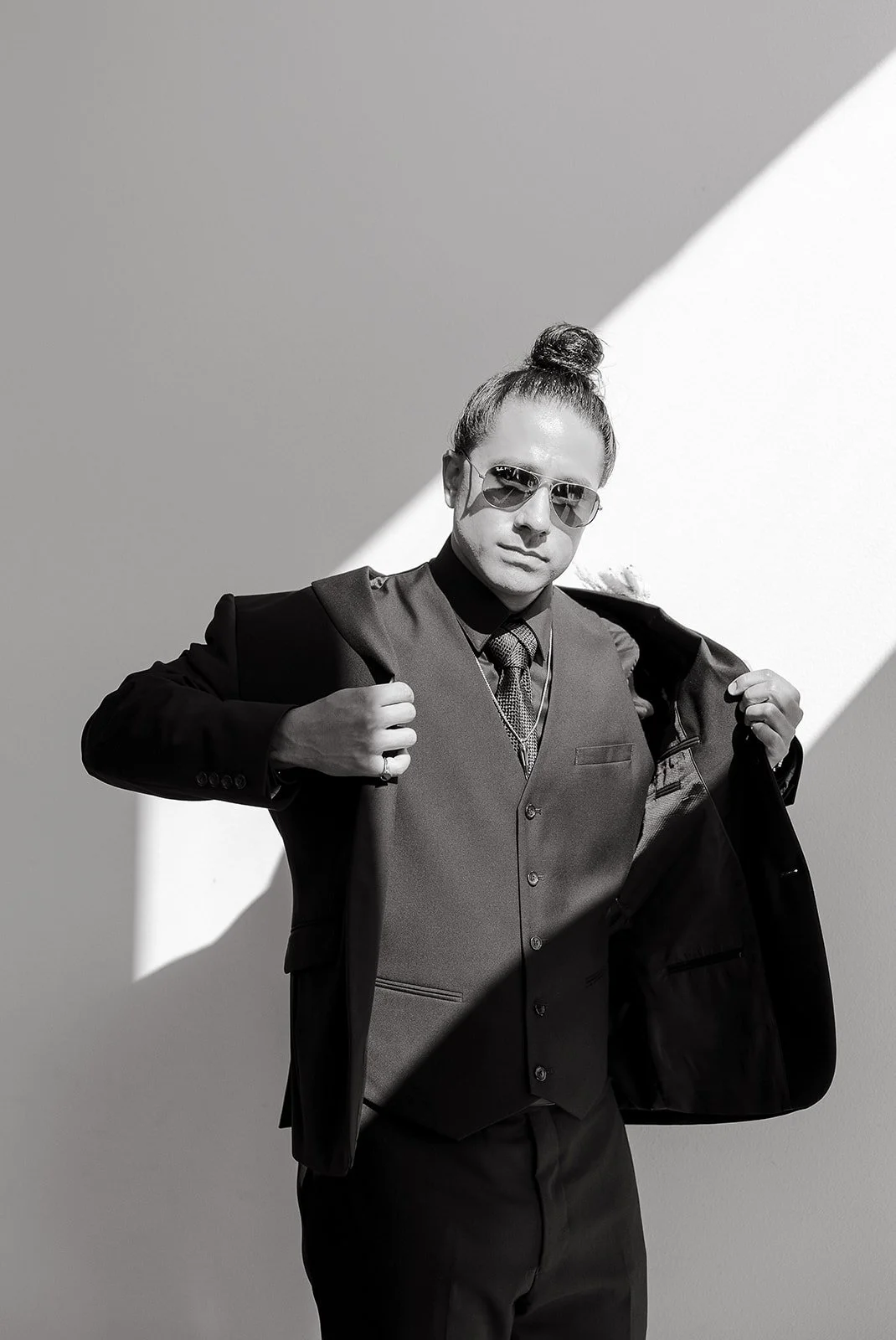 Black and white image of a person wearing sunglasses and a suit, adjusting their jacket under bright sunlight.