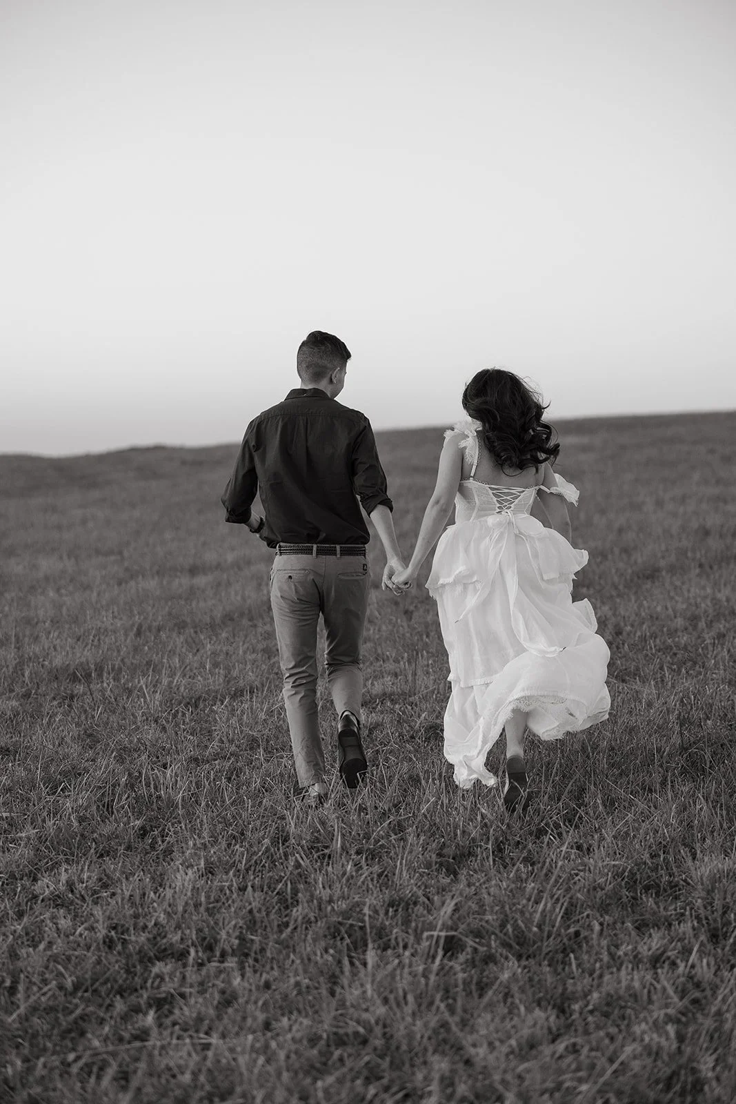 A couple holding hands and walking through a grassy field, with the woman wearing a flowing white dress.